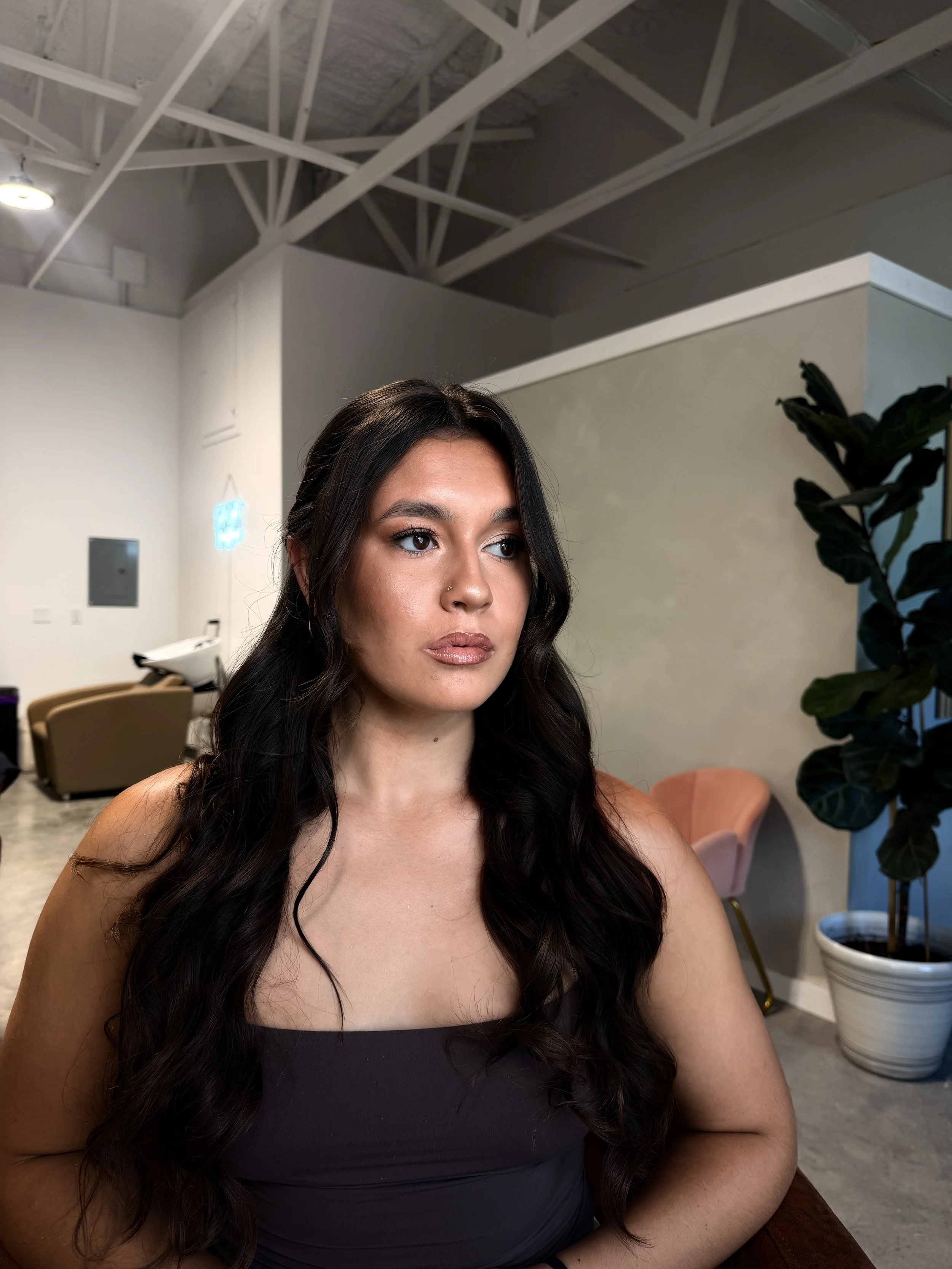 Portrait of a woman with long dark hair, wearing a dark gray strapless top, sitting indoors with a plant and pink chair in the background.
