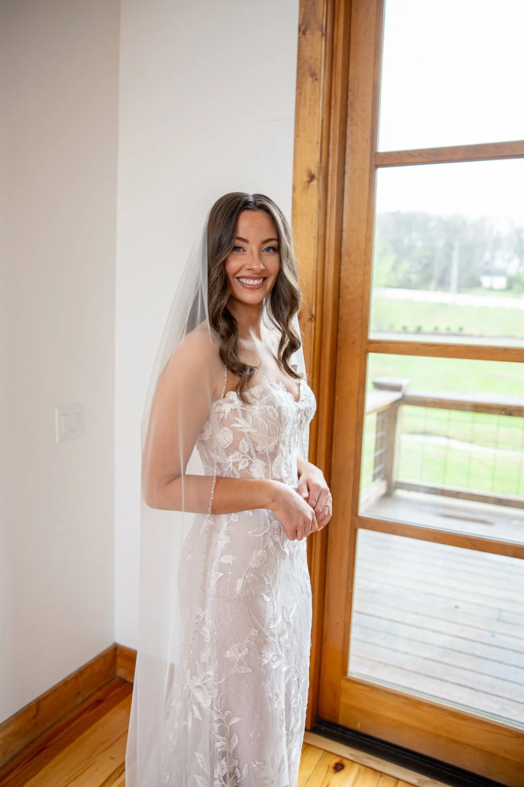 A smiling bride standing by a wooden door in a lace wedding dress with a veil, holding her hands in front of her, in a room with wood trim and a view of a green outdoor area.