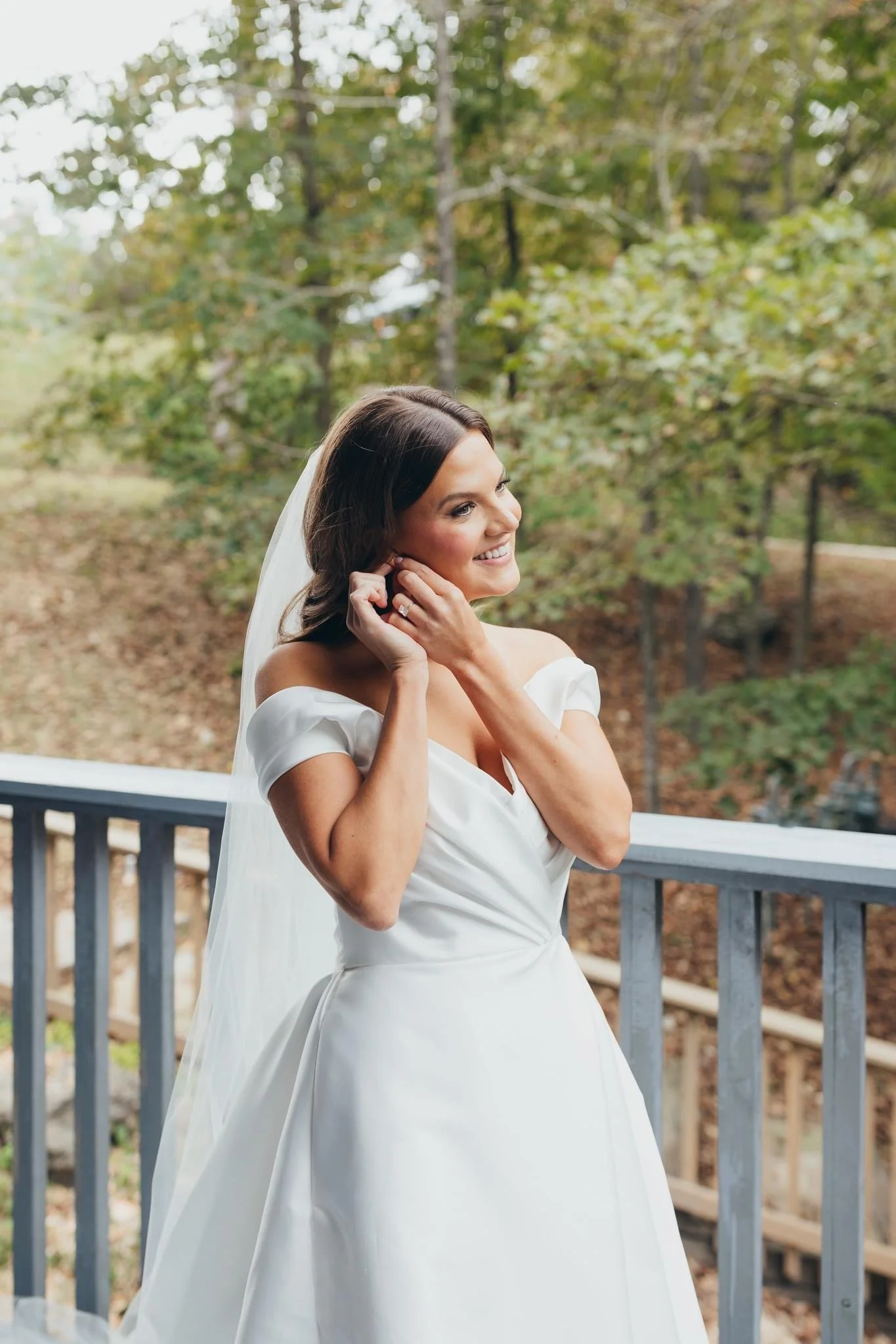 A woman in a white wedding dress standing outdoors on a wooden deck, smiling and adjusting her earring with trees and foliage in the background.
