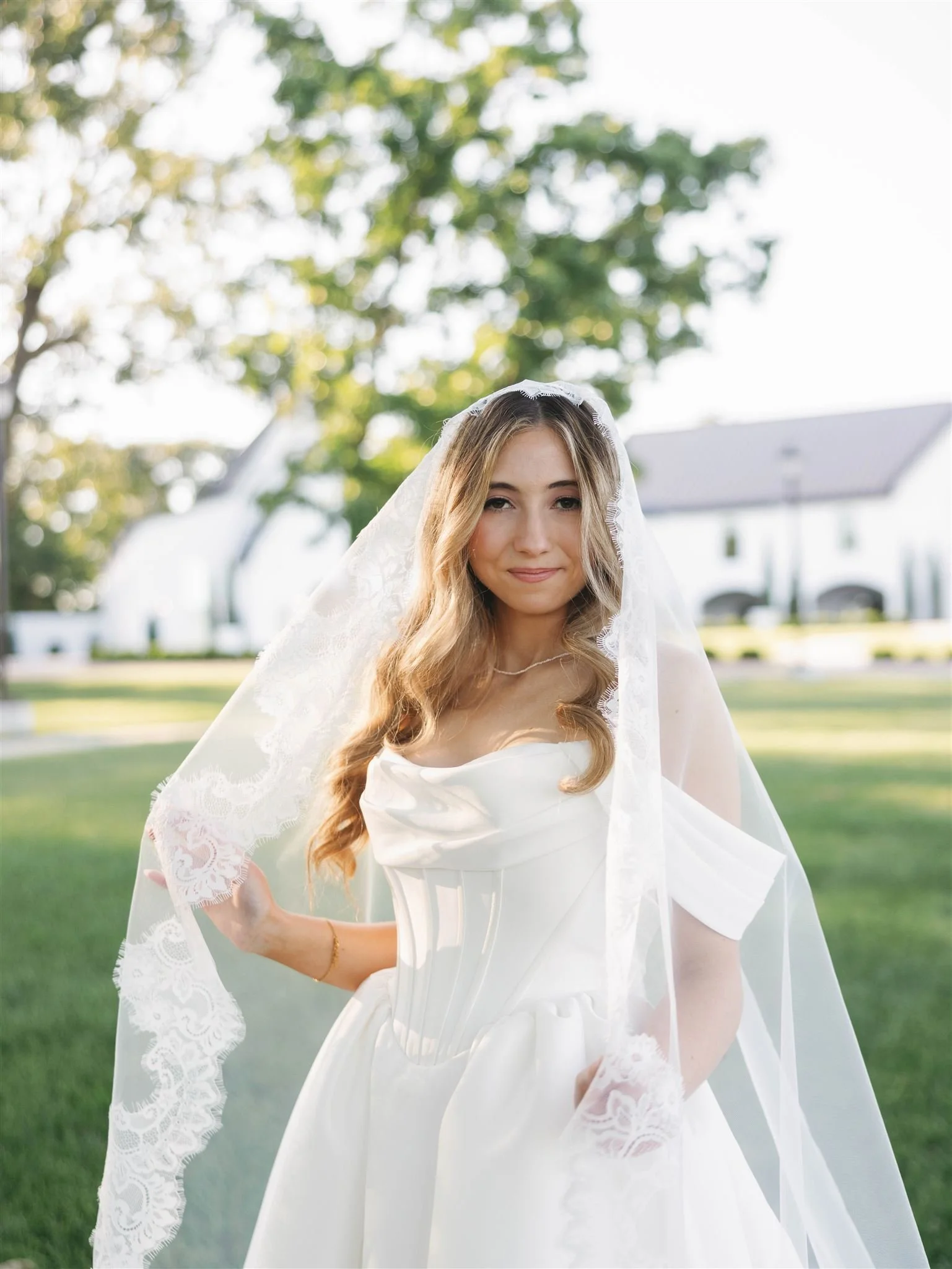 A young woman in a wedding dress and veil stands outdoors on a sunny day, with trees and a white house visible in the background.