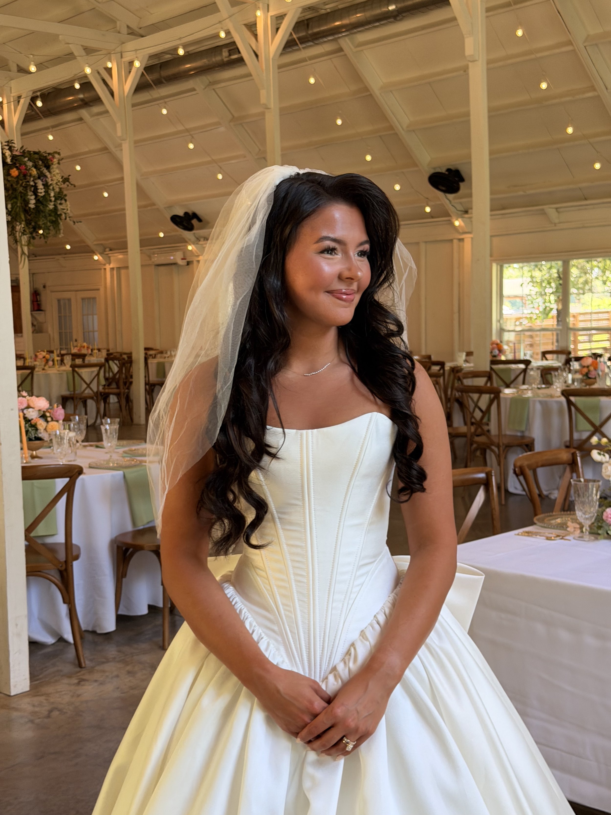 A woman dressed in a wedding gown with a veil is standing inside a decorated reception hall. The hall has string lights hanging from the ceiling, tables with floral centerpieces, and chairs arranged around the tables.