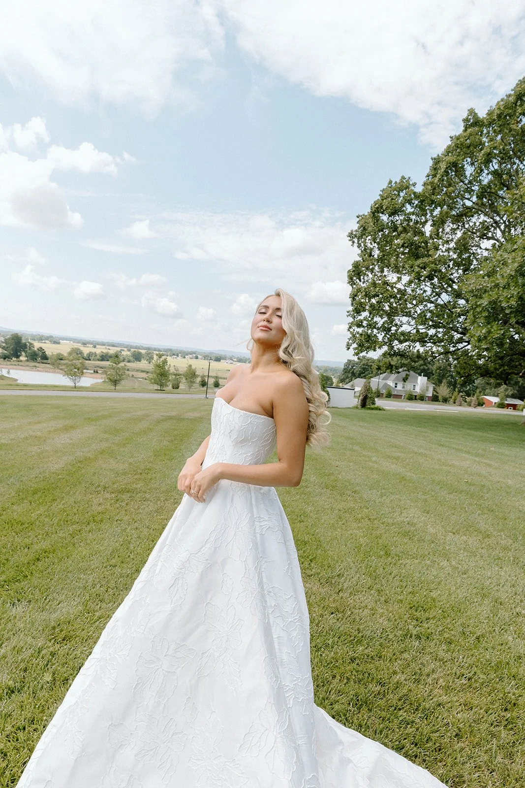 A woman in a white strapless wedding dress standing on a grassy field with trees and a blue sky with clouds in the background.