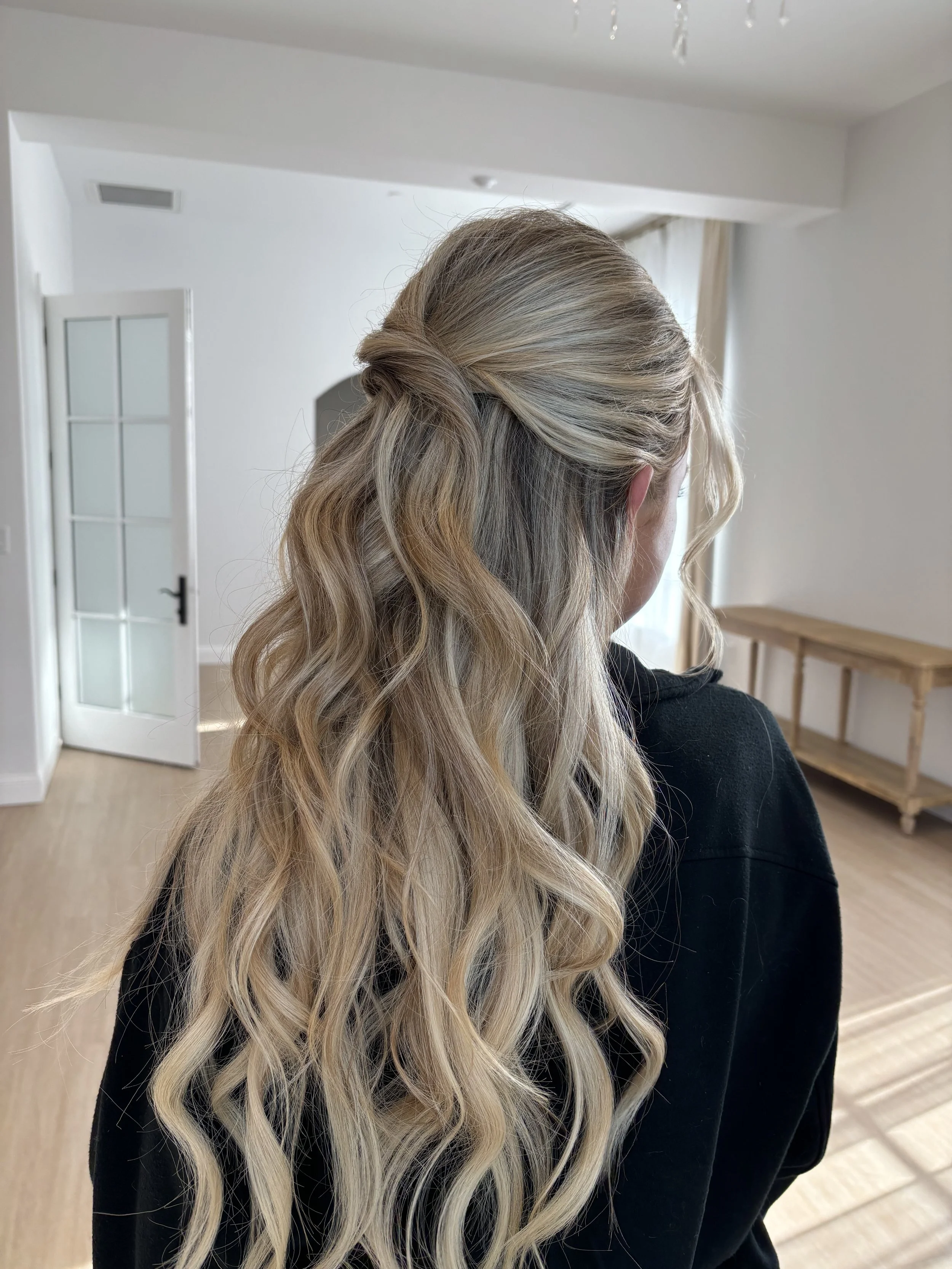 A woman with long, wavy blonde hair styled in a half-up, half-down hairstyle standing in a bright room with white walls and wooden flooring.