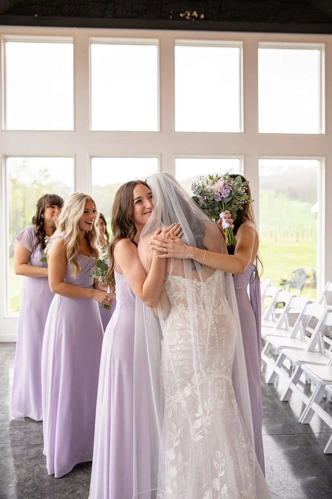 A bride in a white lace wedding gown is hugging a bridesmaid in a lavender dress, who is holding a bouquet of flowers, while other bridesmaids wait behind them, all smiling and sharing a joyful moment in a bright room with large windows.