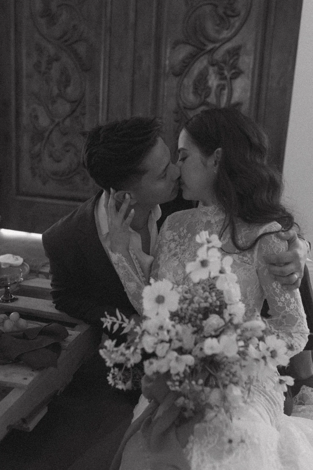 A black and white photo of a couple kissing at their wedding. The woman wears a lace wedding dress and holds a bouquet of flowers, while the man is in a suit. They are embracing in front of a decorative wooden wall.