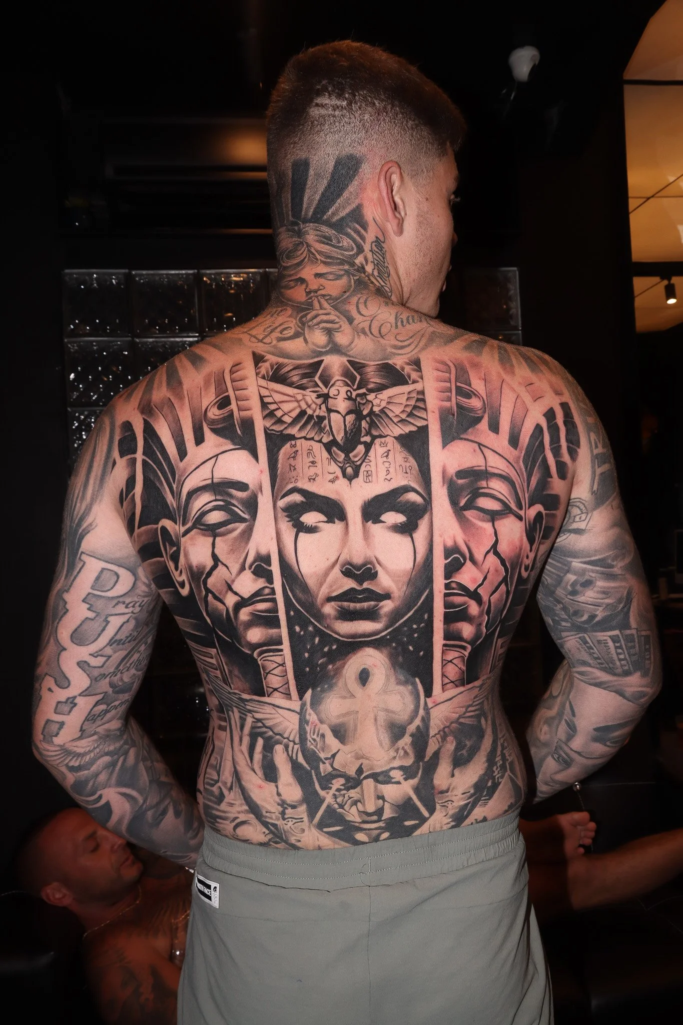 Man with extensive black and gray tattoos covering his back and arms, featuring faces, symbols, and intricate designs in a monochrome style.