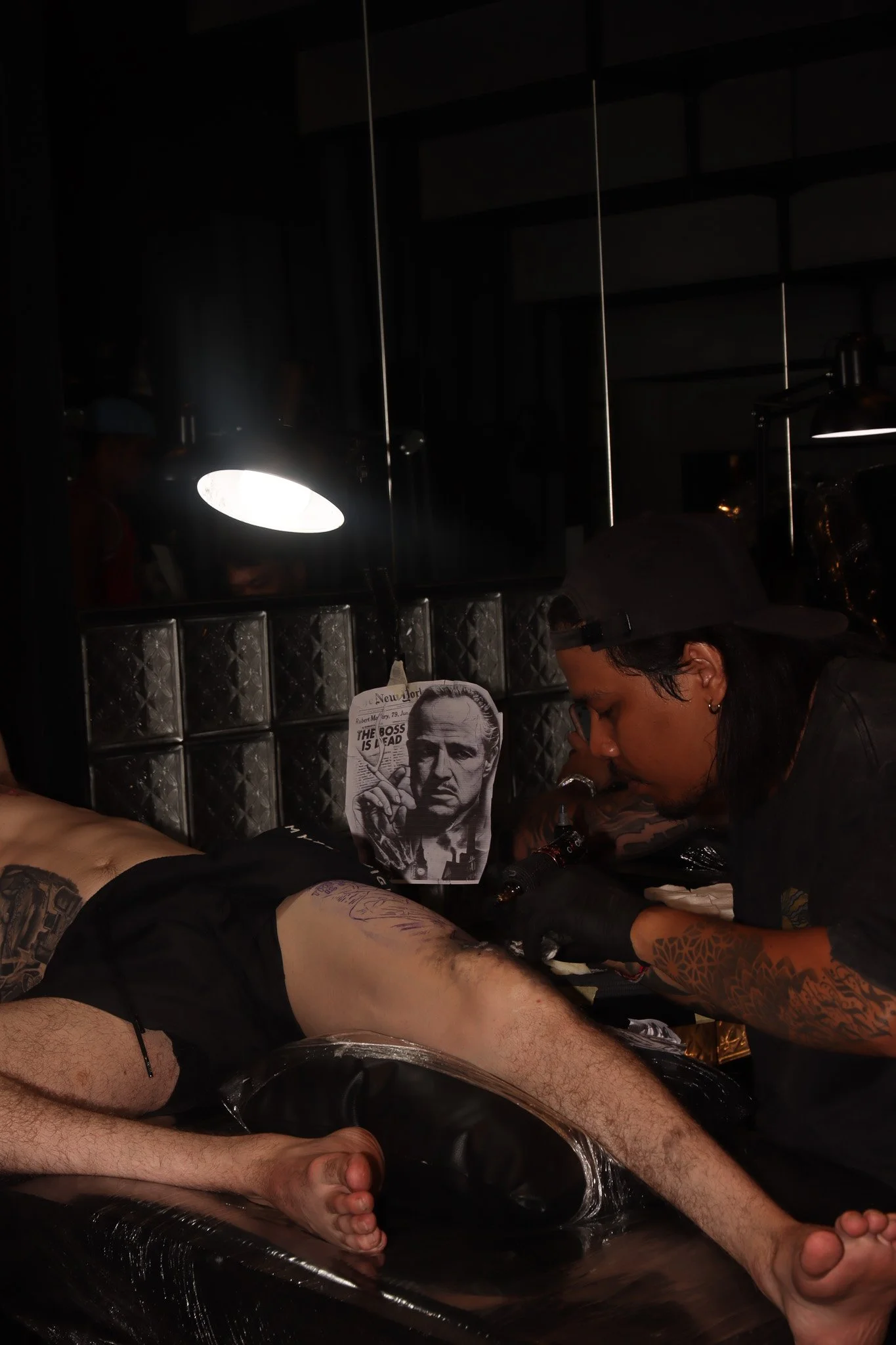 A tattoo artist working on a man's leg while lying on a black tattoo table in a dimly lit environment, with a black-and-white cutting of a man's face in the background.