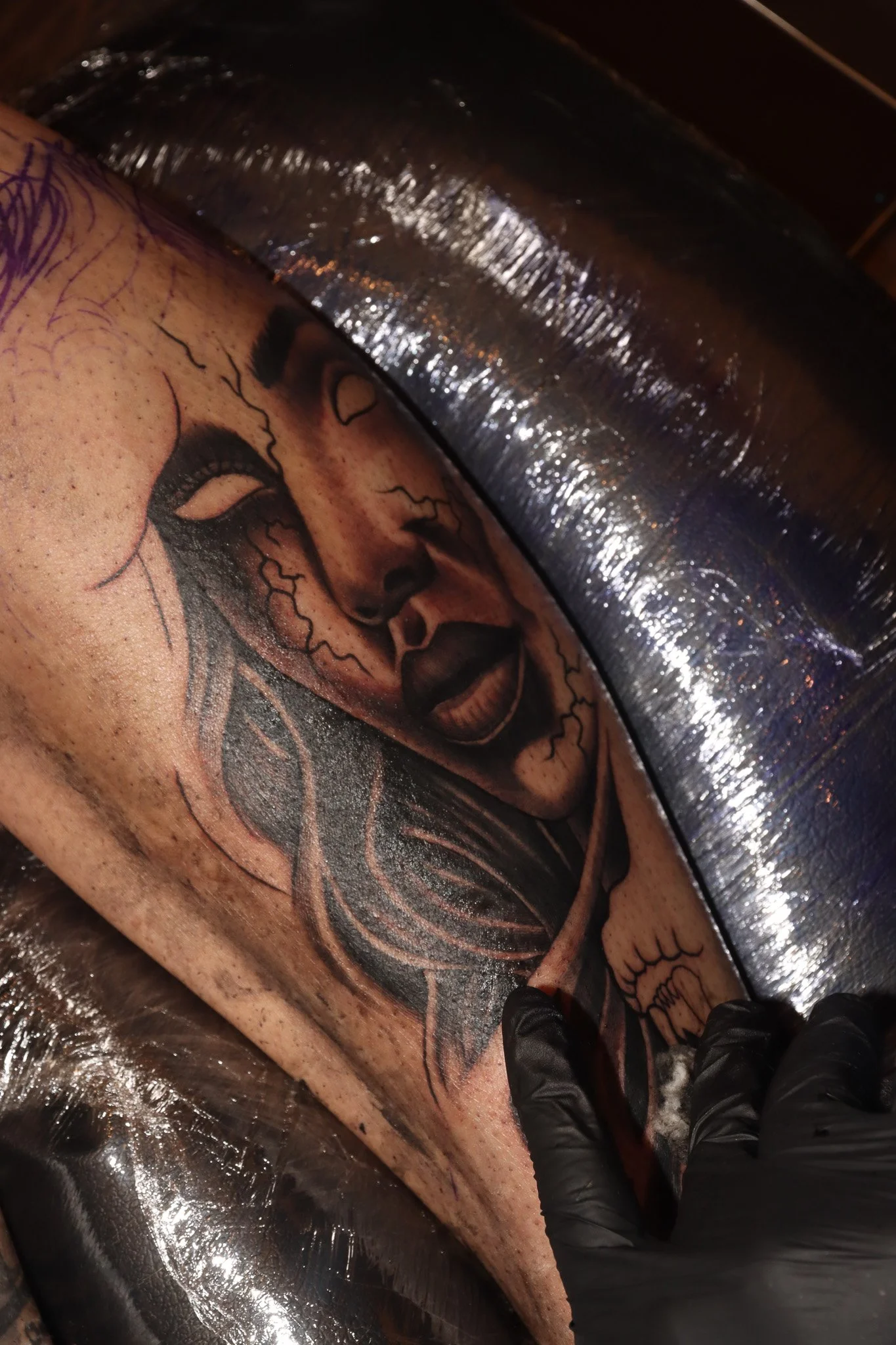 A tattoo artist is creating a detailed black and gray tattoo of a woman's face with long hair, closed eyes, and a peaceful expression on a person's arm, with the arm covered in skin pigment and the person wearing black gloves.