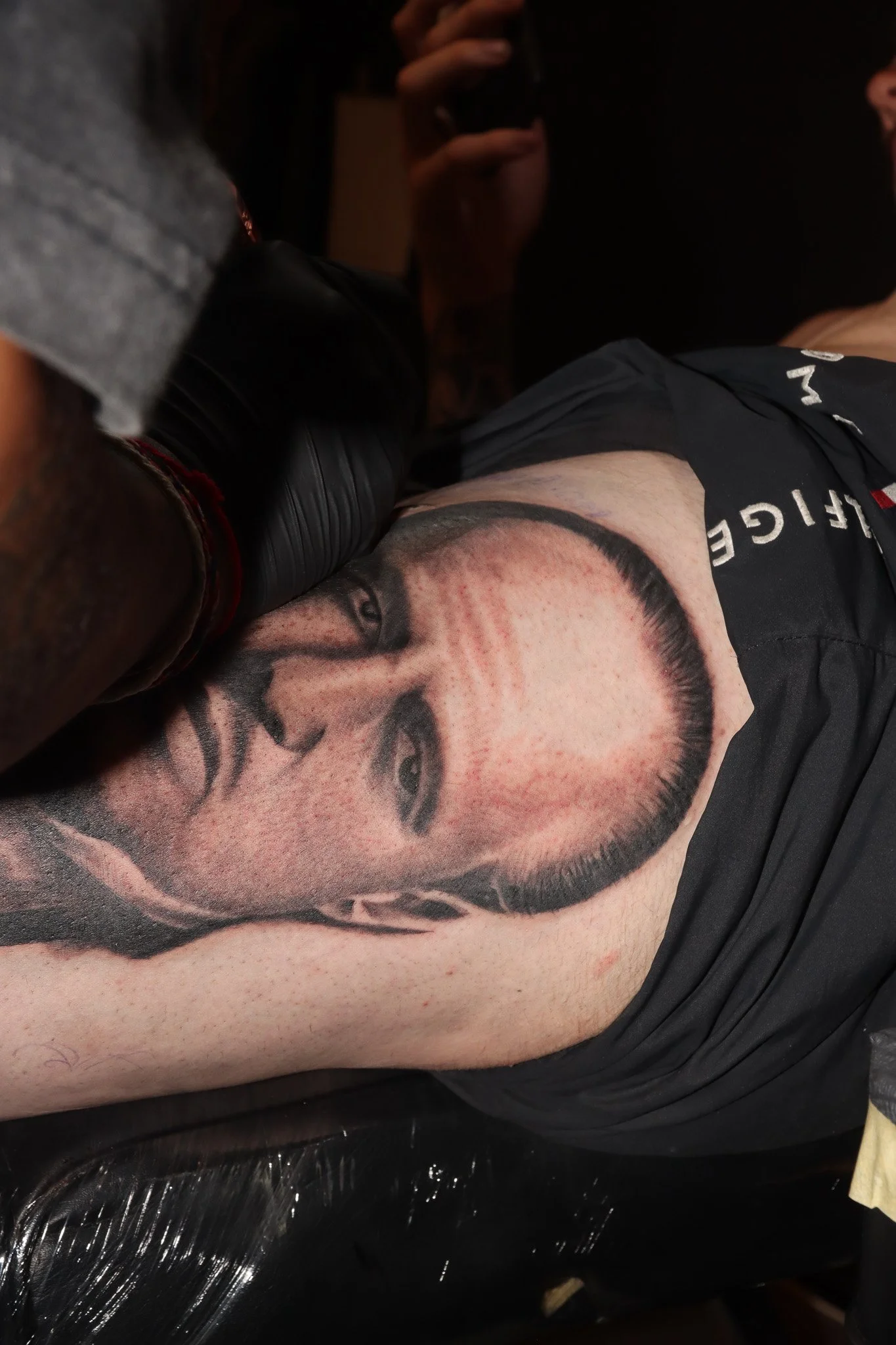 A person receiving a tattoo of a woman's face on their upper arm or shoulder. The tattoo appears to be black and gray, depicting a realistic portrait. The person is lying down, wearing a black shirt, and a tattoo artist's gloved hand is visible worki