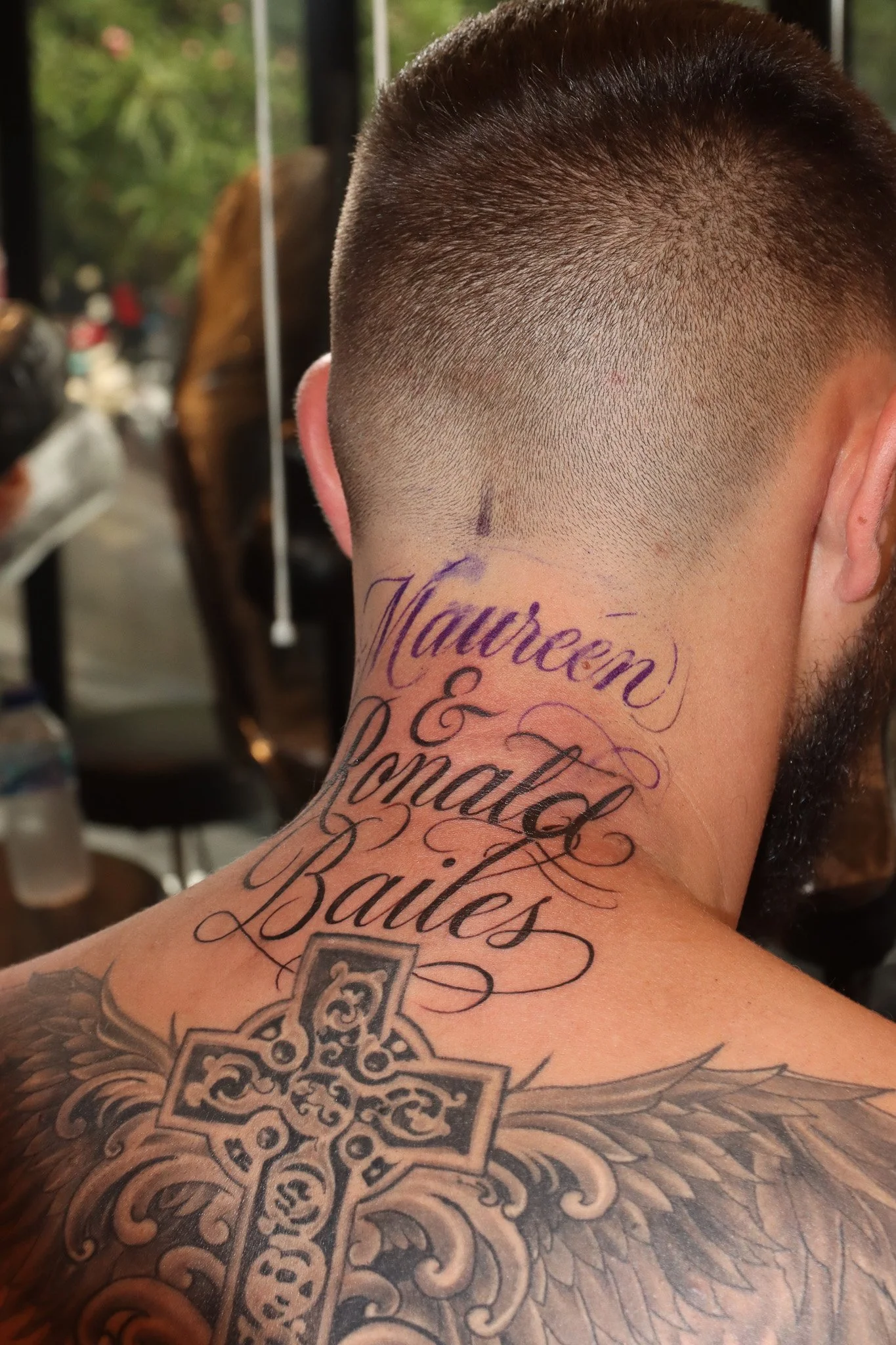 Tattoo on the back of a man's neck that reads 'Nouwen & Ronald Baile' in cursive script, with a detailed Celtic cross and feather design on his shoulder.
