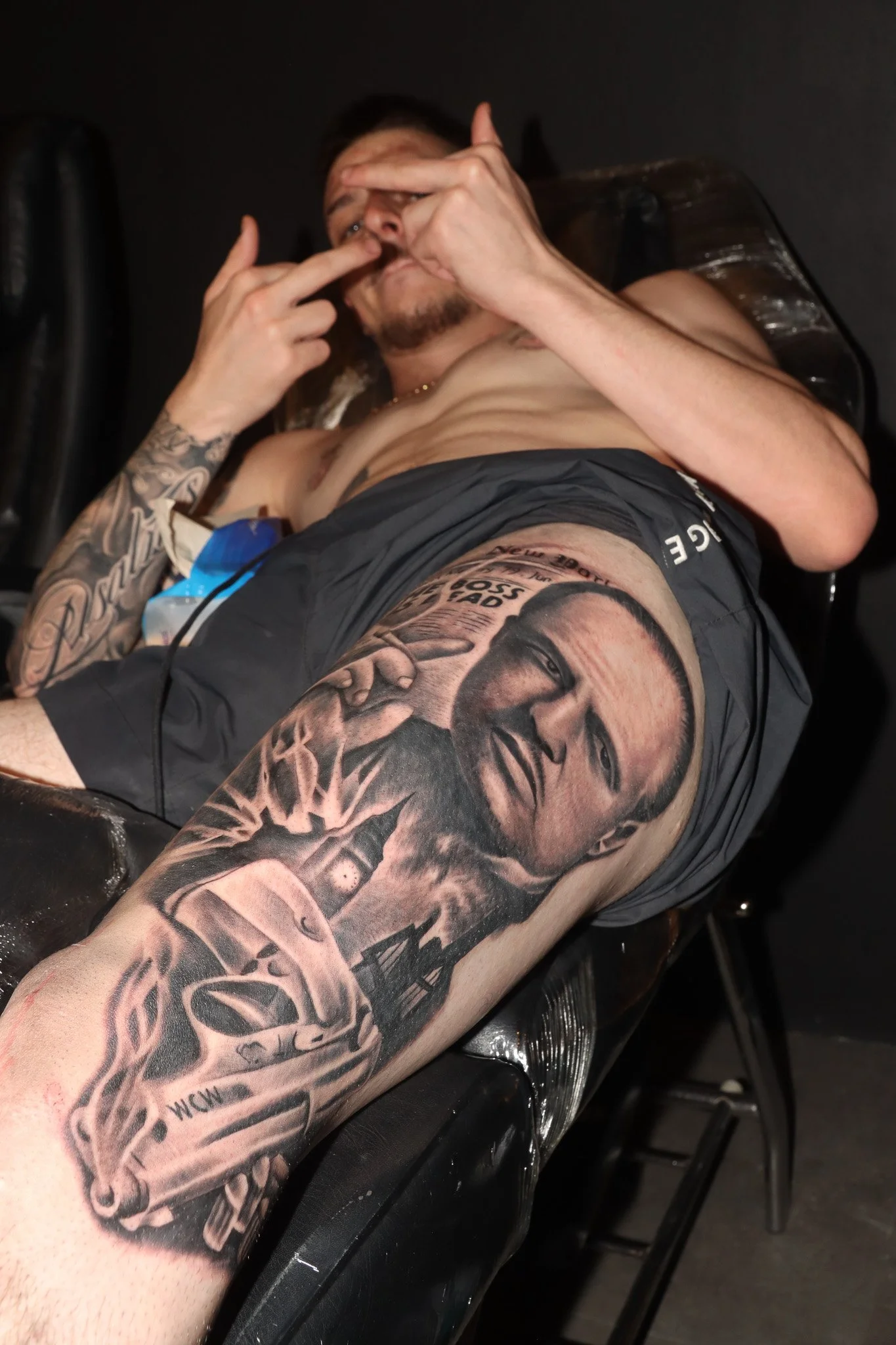 A shirtless man with tattoos on his arm and leg, lying on a chair or bed, making a hand gesture and pointing to his forehead, with a dark background.