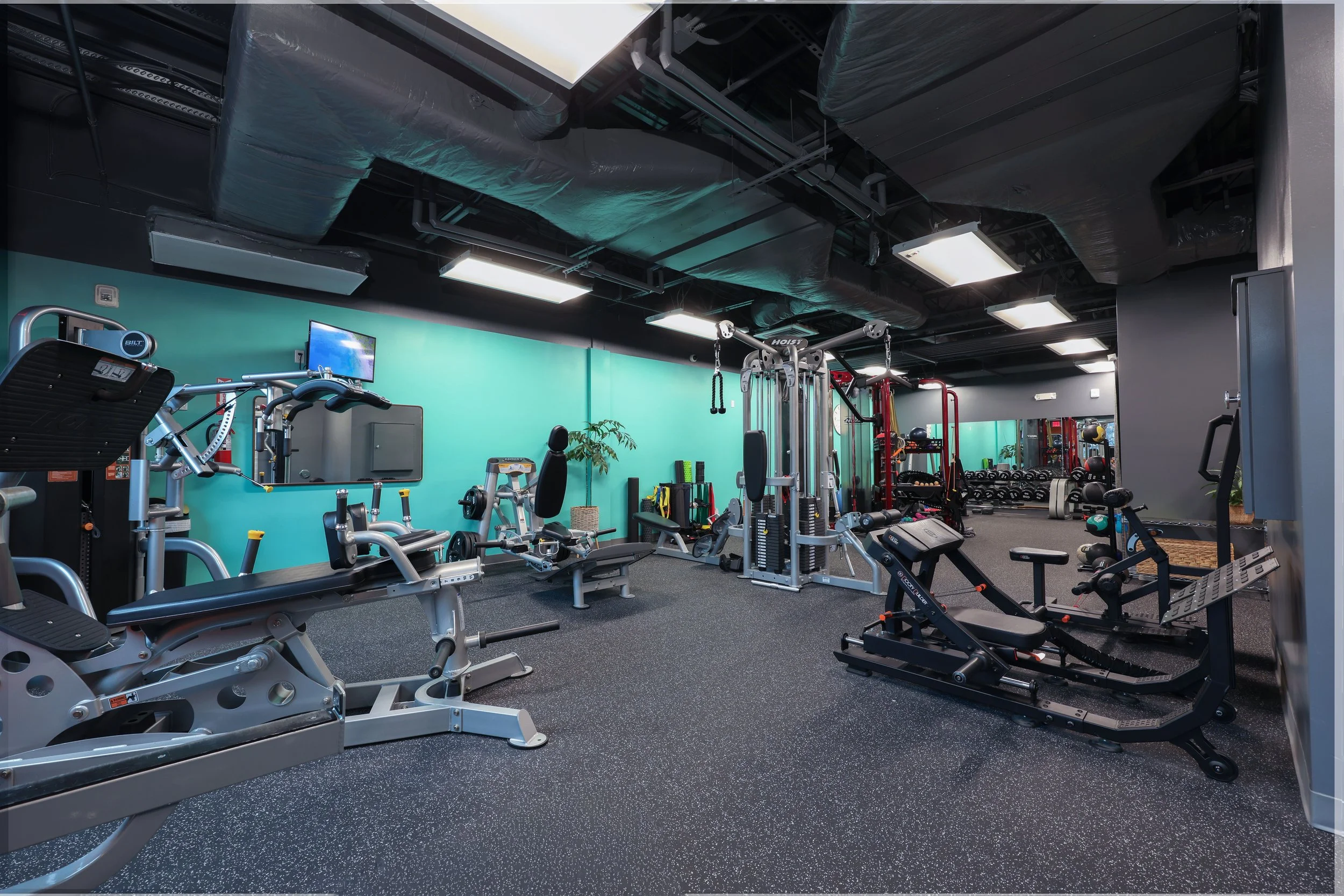 Indoor gym with various workout equipment including exercise bikes, weight machines, a rowing machine, and free weights against a teal wall ceiling with black painted ducts.