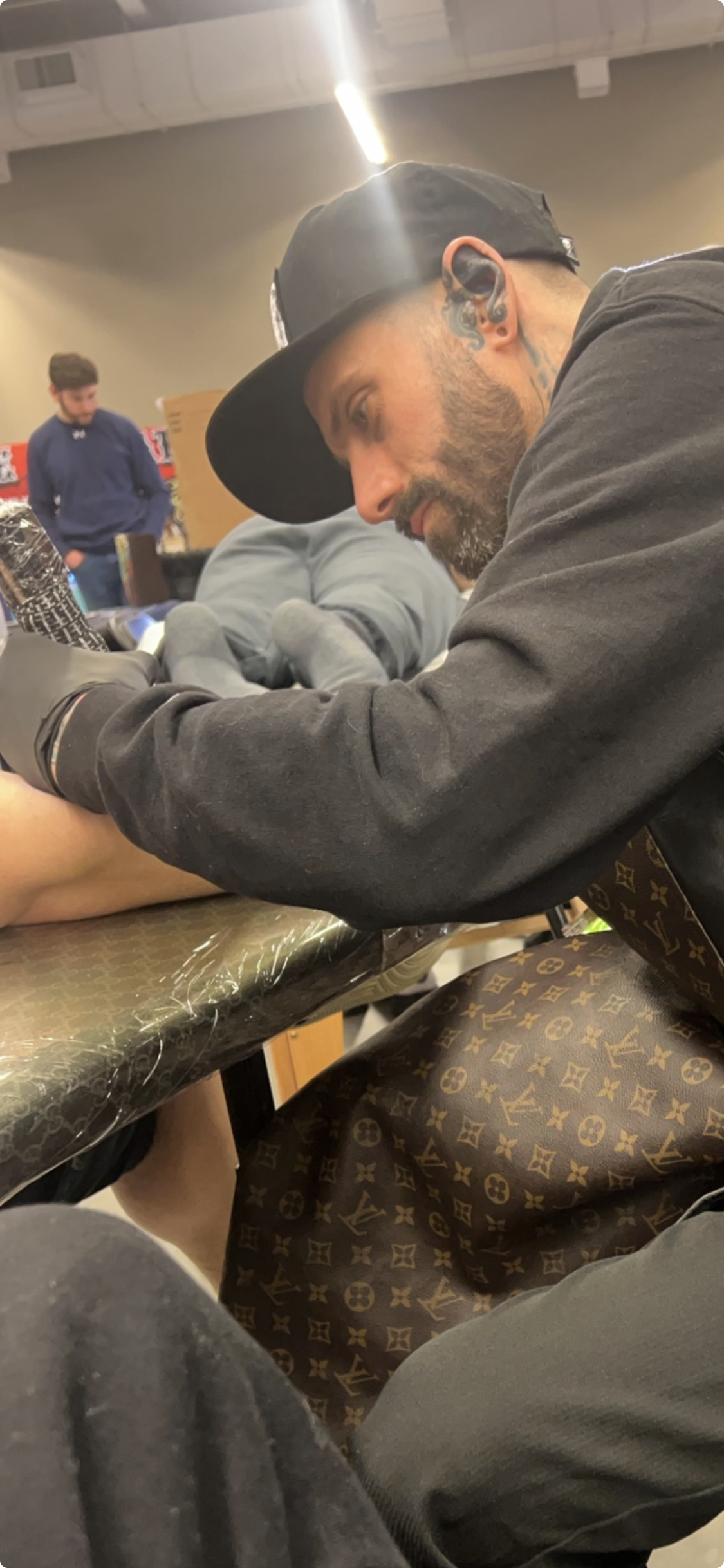 Tattooed man with a beard and a black cap getting a tattoo on his foot, with another person lying on a table in the background.