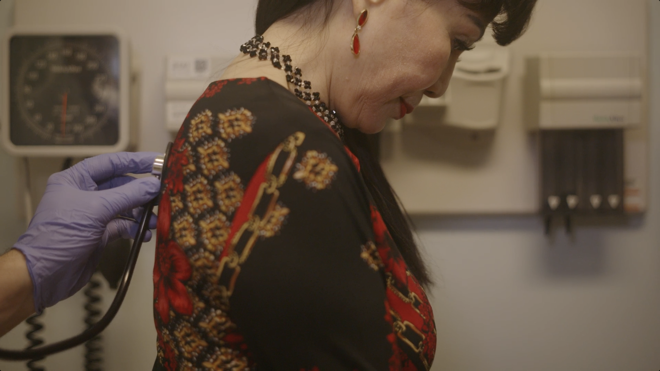 A healthcare professional in purple gloves uses a stethoscope on an Asian woman wearing a black dress with red, gold, and orange embroidery, in a clinical setting.