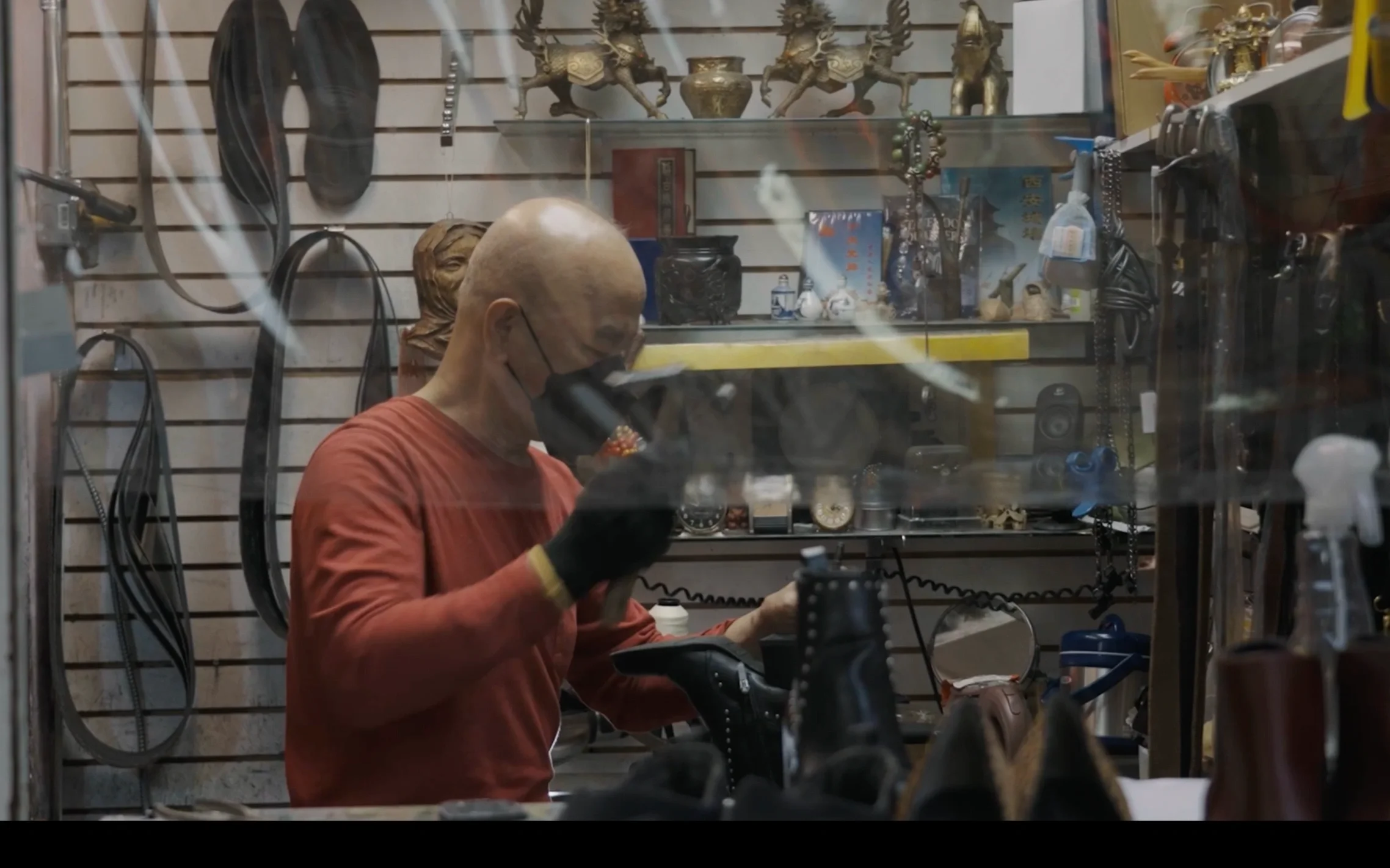 A person with a bald head, wearing a face mask and gloves, working inside a shop with various tools and old clocks displayed on walls and shelves.