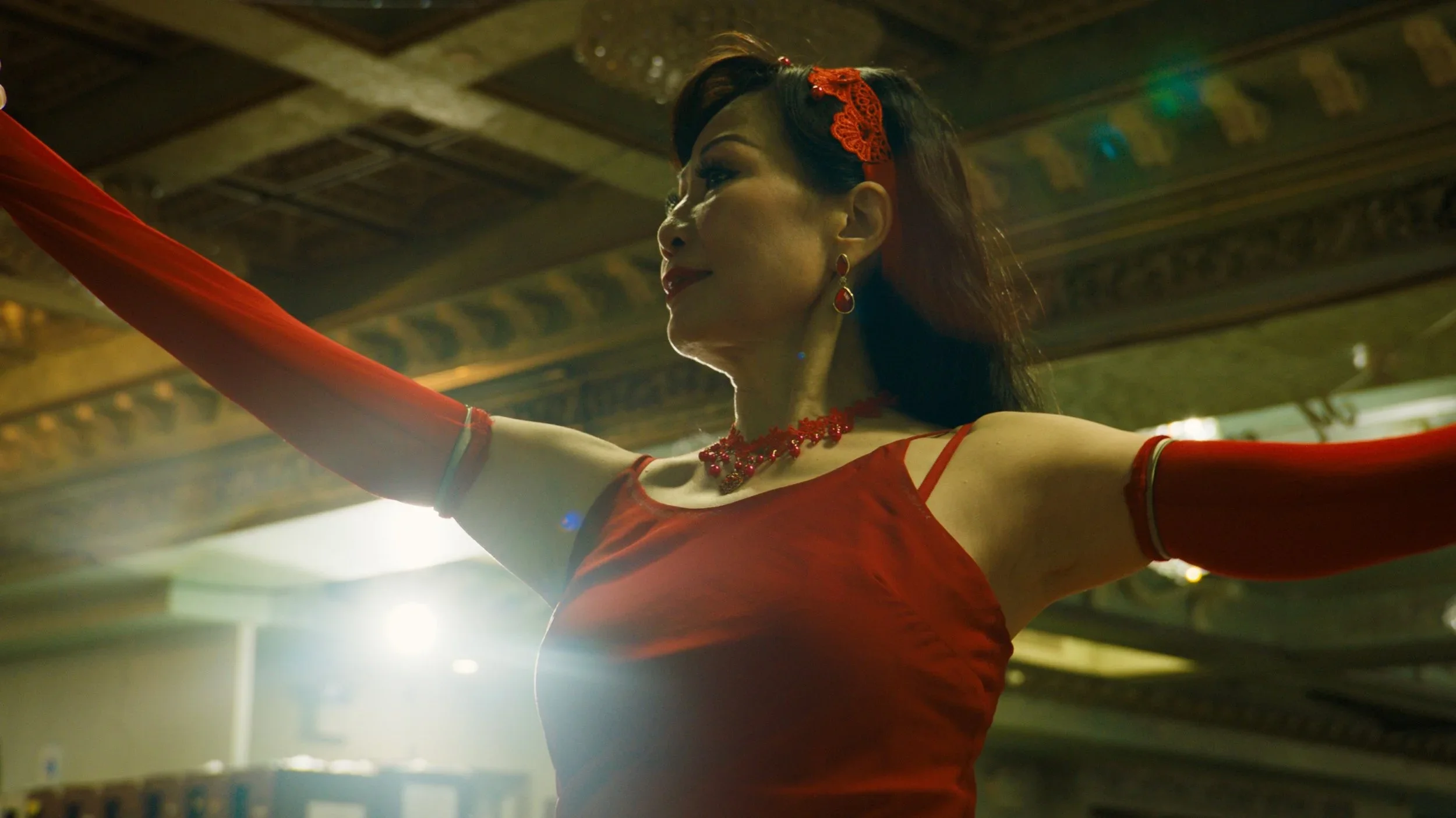A woman in a red dress with matching gloves and jewelry, posing with arms outstretched under ornate ceiling decor.