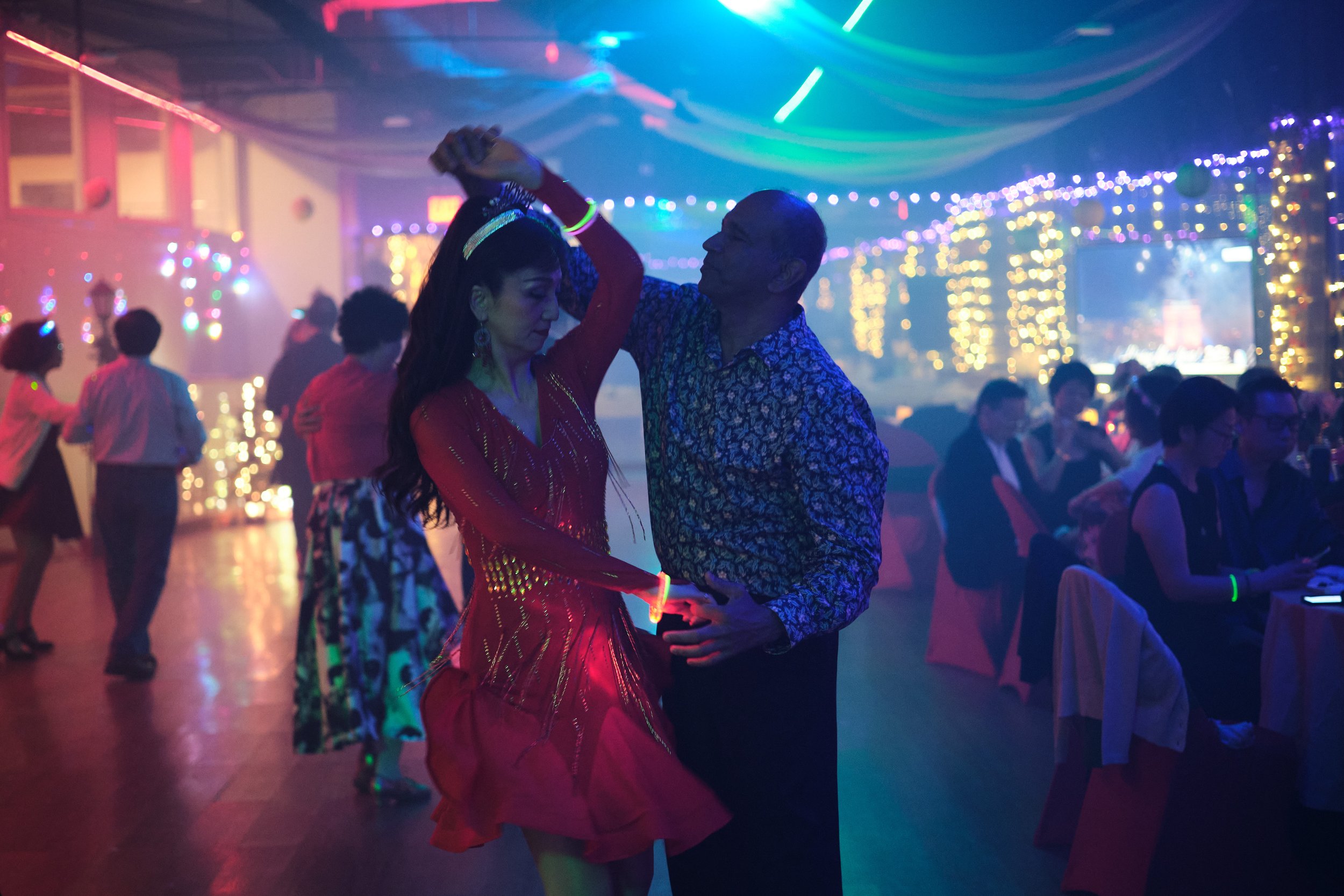 Couple dancing at a party with colorful lights and decorations, other guests sitting and dancing in the background.