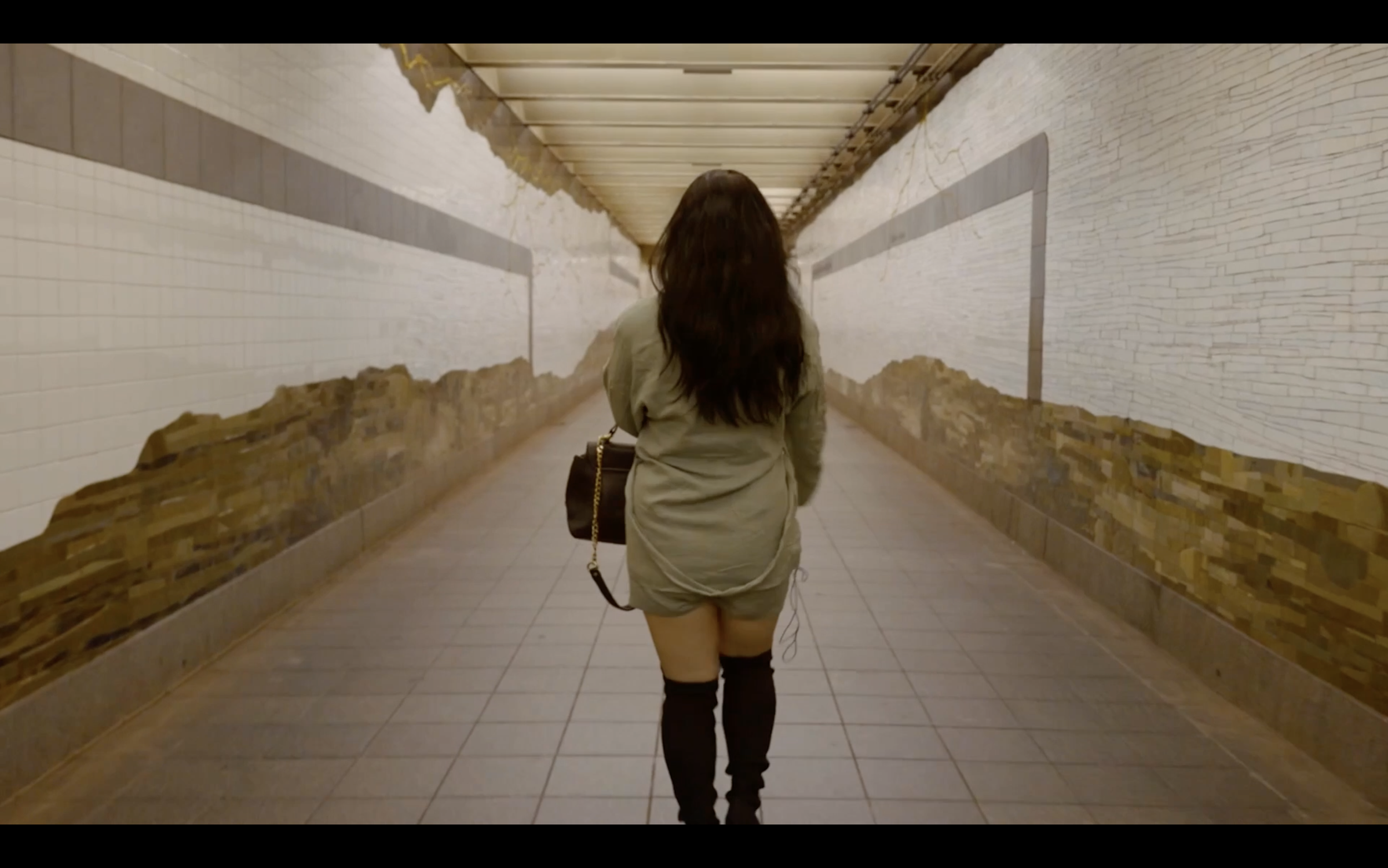 A woman with long dark hair from behind walking through an underpass tunnel with white walls and a tiled floor.