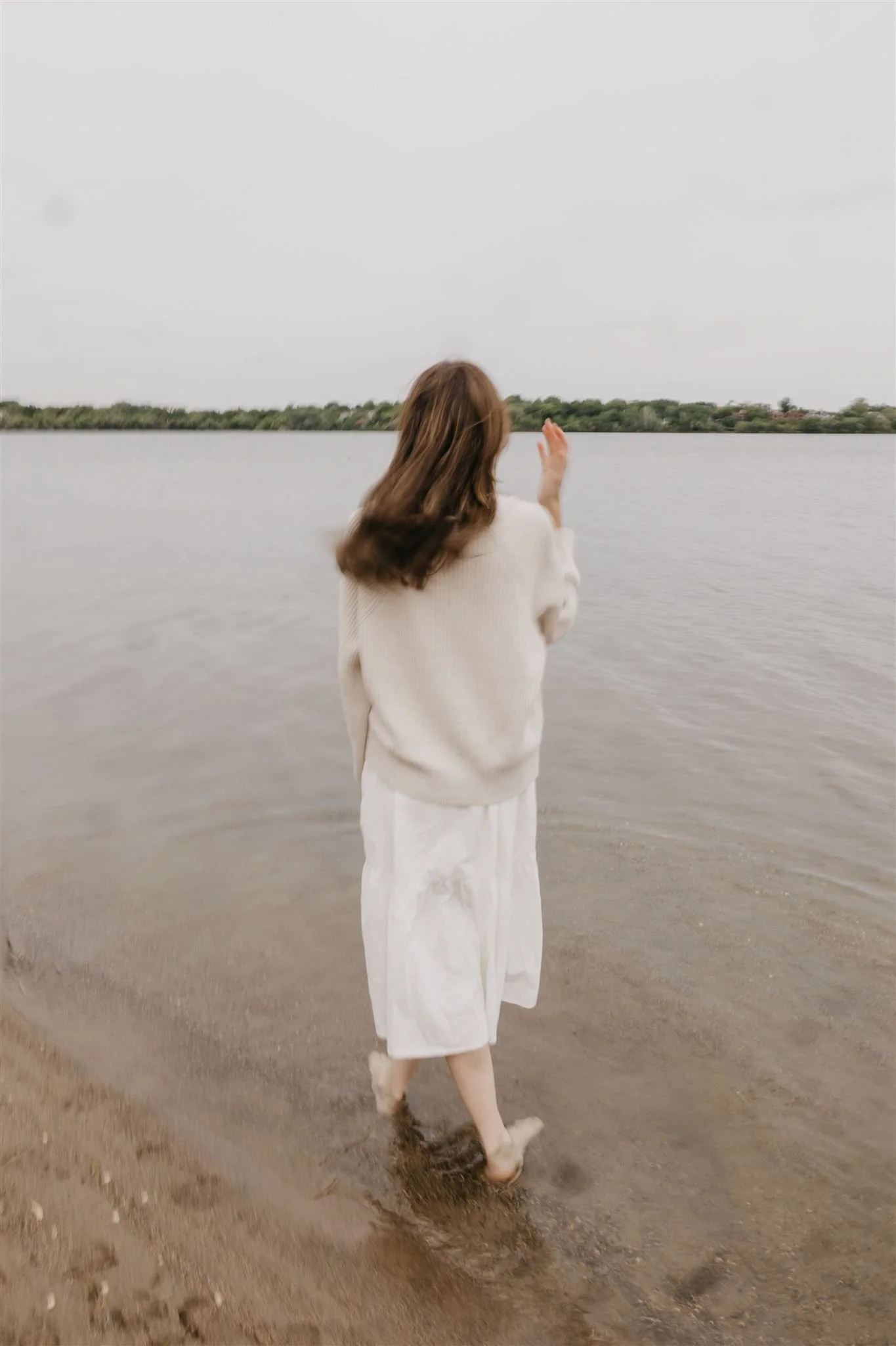 A woman standing at the edge of a lake with her back to the camera, wearing a beige sweater and white skirt, with her feet in the water and looking out at the lake.