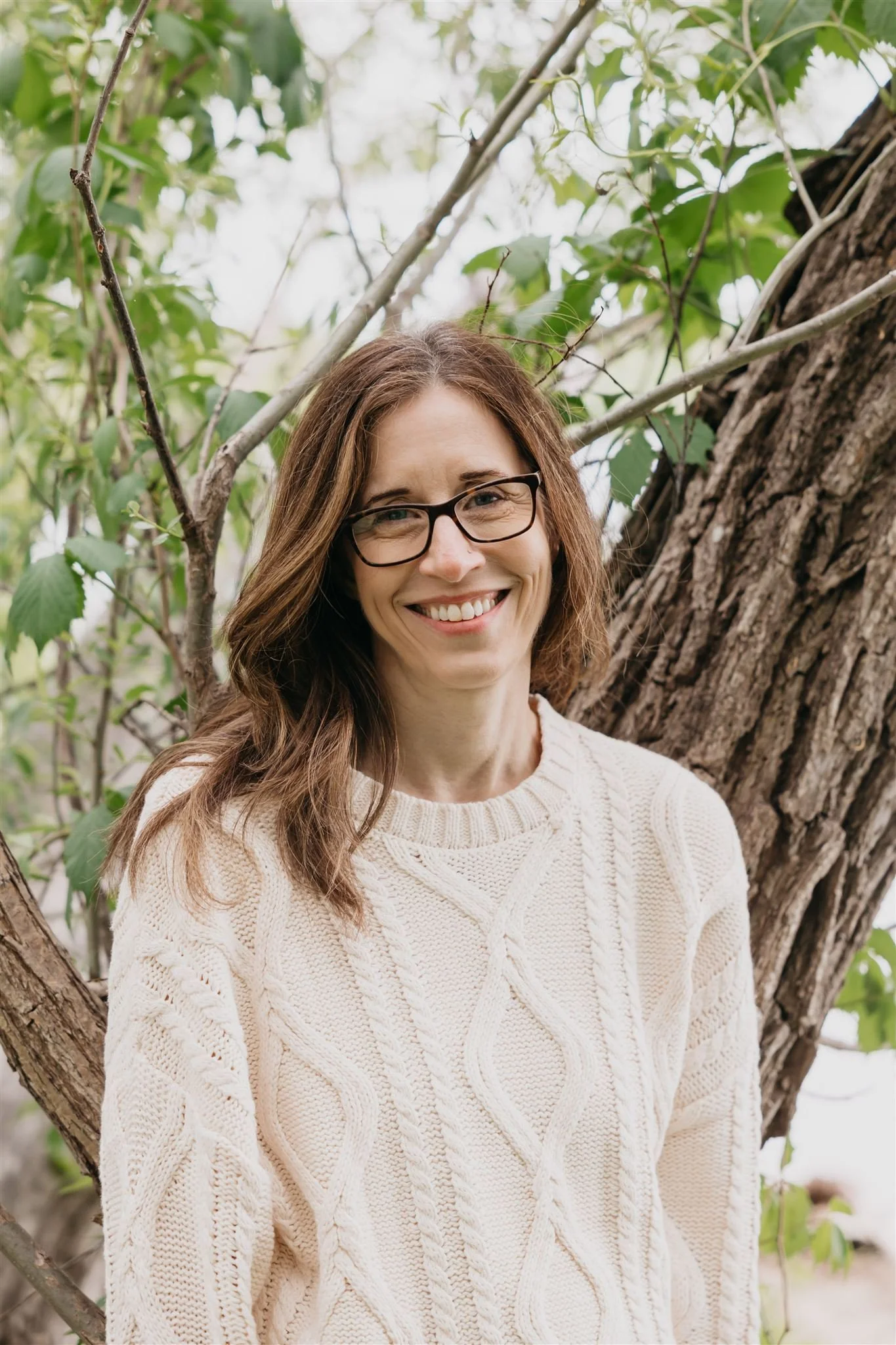 A woman with brown hair, glasses, and a cream-colored knitted sweater smiling outdoors near a tree with green leaves.