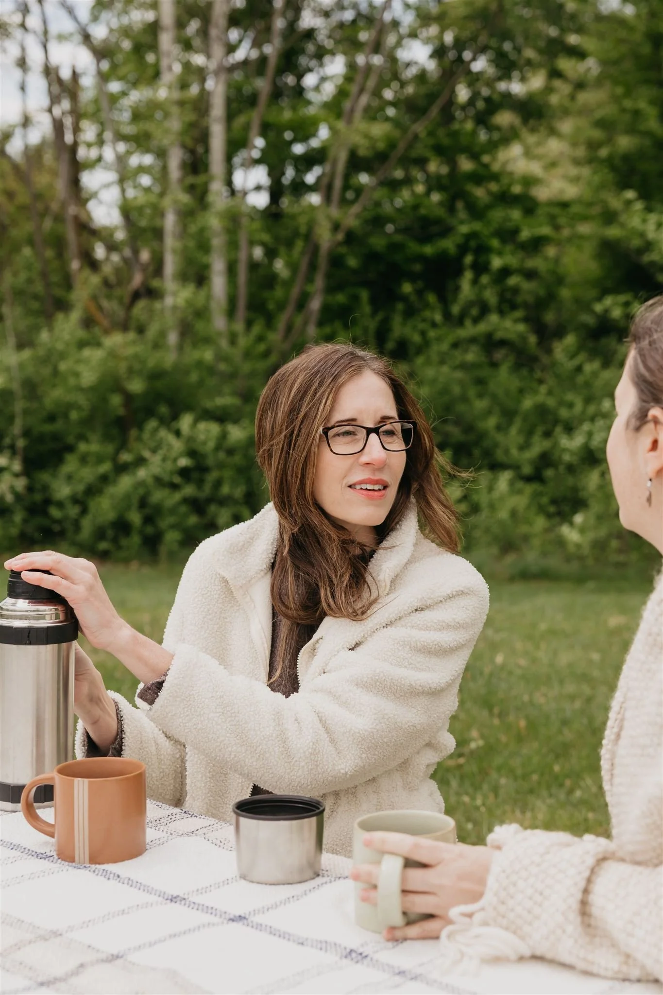 Two women having coffee outdoors at a picnic table with mugs, a thermos, and a lunch box, surrounded by green trees.