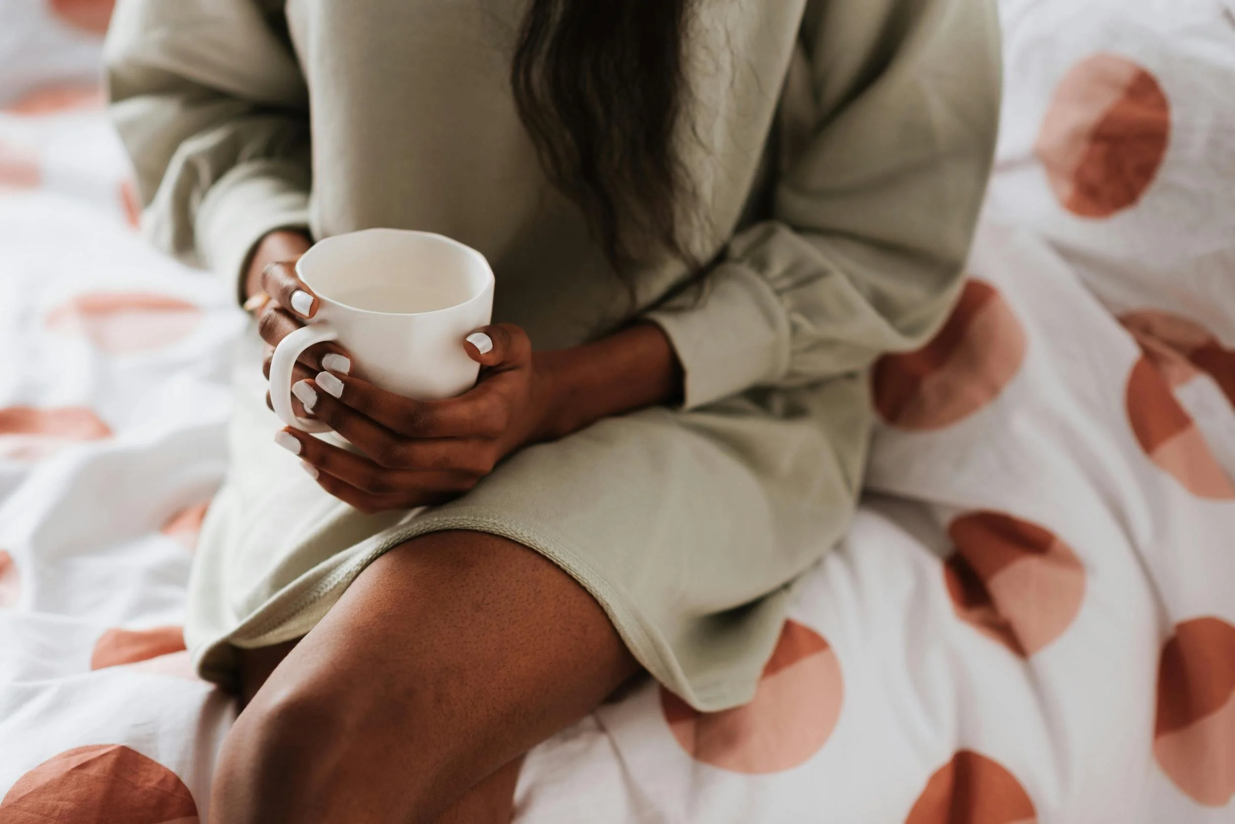 Person sitting on bed holding a white mug, wearing a beige outfit with a polka dot bedspread.