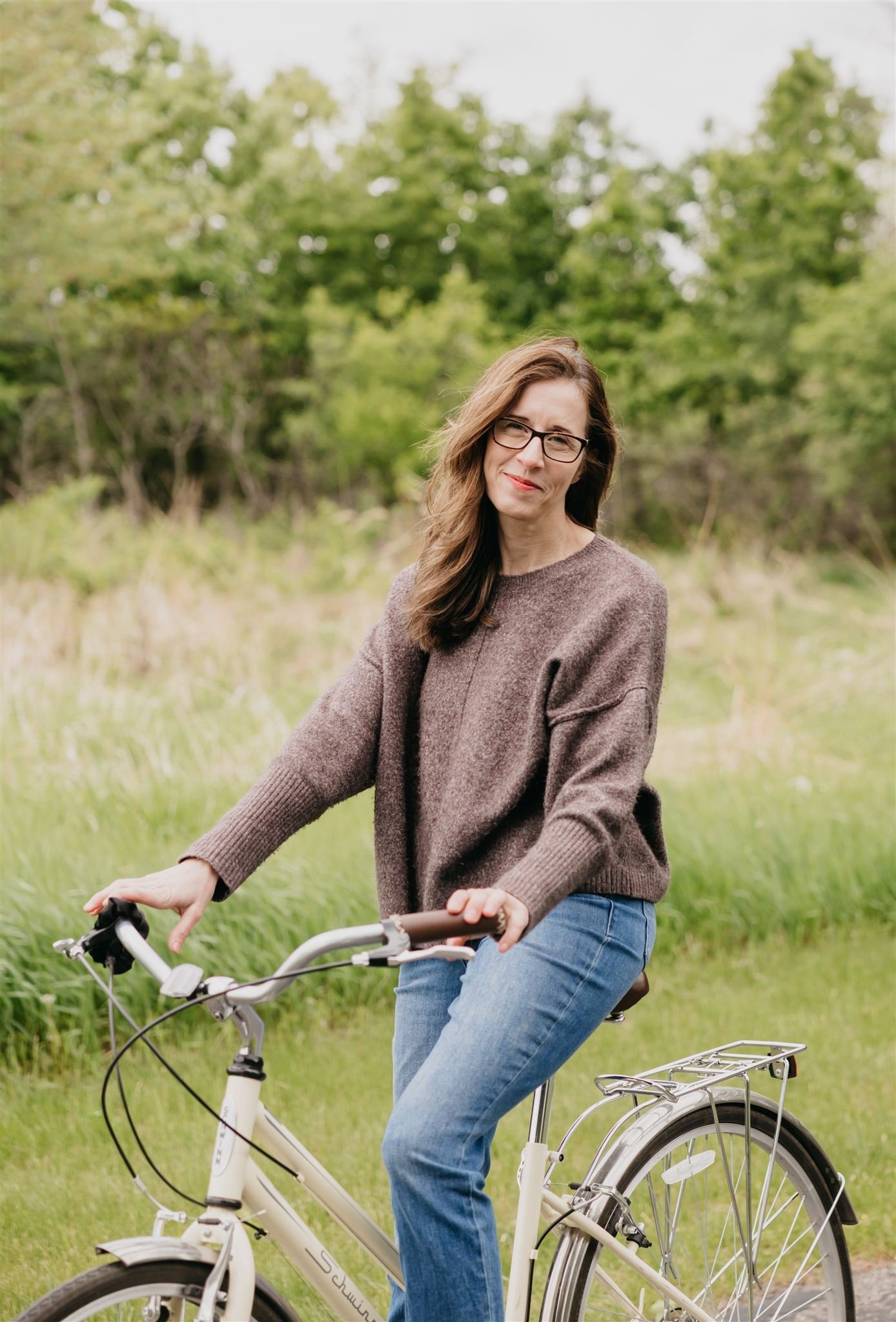 A woman with long brown hair and glasses riding a white bicycle outdoors in a grassy area with trees in the background.