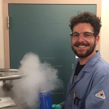 A man with curly hair and glasses wearing a blue lab coat and green gloves, smiling in a laboratory with a green chalkboard behind him. Smoke or vapor is rising from a container in front of him.