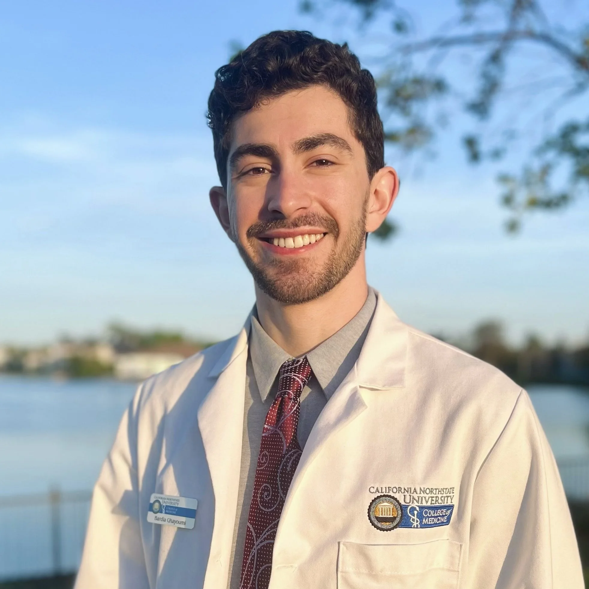 Young man in a white lab coat smiling outdoors with water and trees in the background, wearing a name tag and medical school patches.