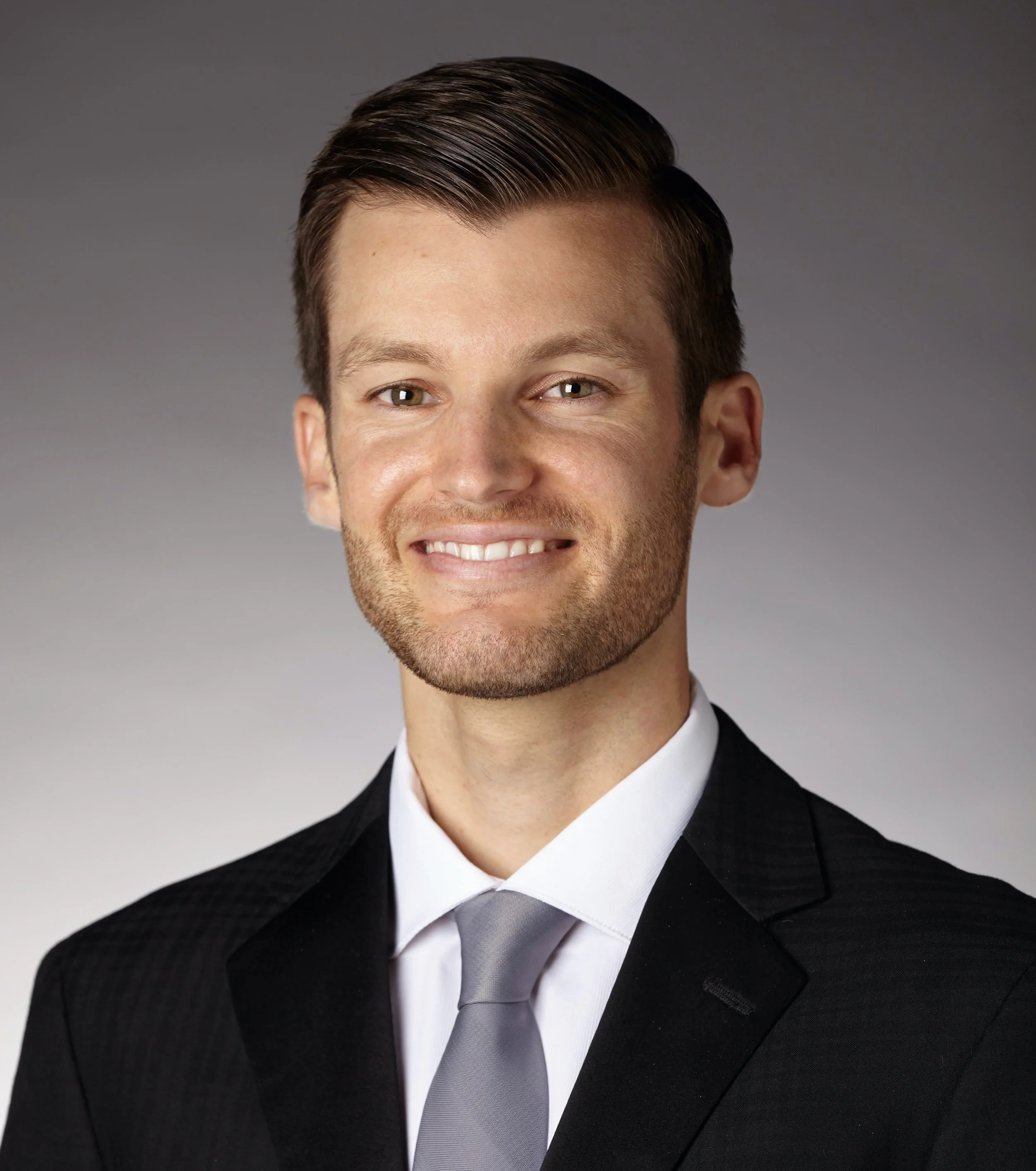 Professional headshot of a smiling young man with brown hair, wearing a black suit, white shirt, and gray tie, against a neutral background.