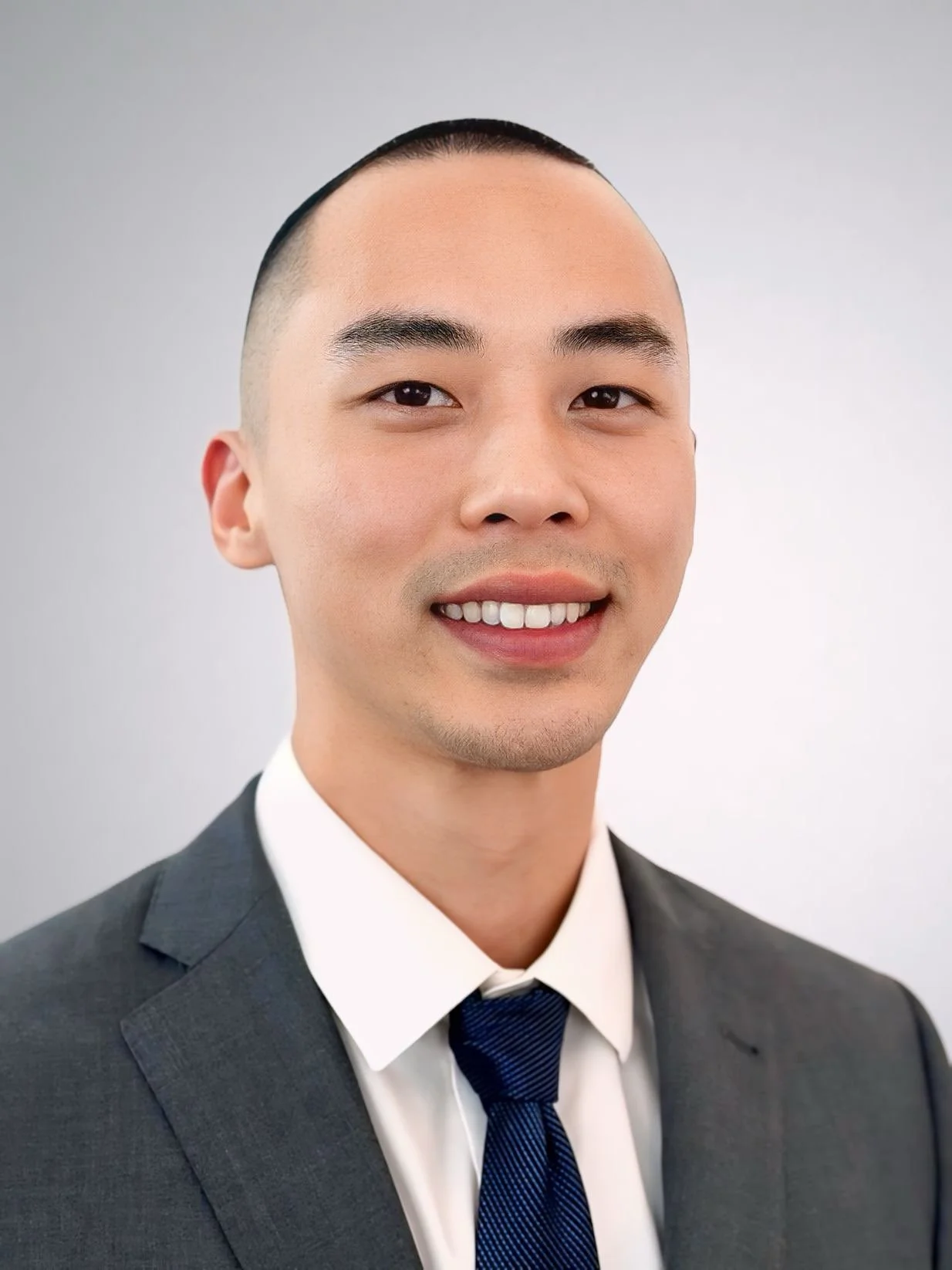 Professional headshot of a smiling young Asian man with short hair, wearing a dark suit, white shirt, and dark blue striped tie, against a plain light gray background.