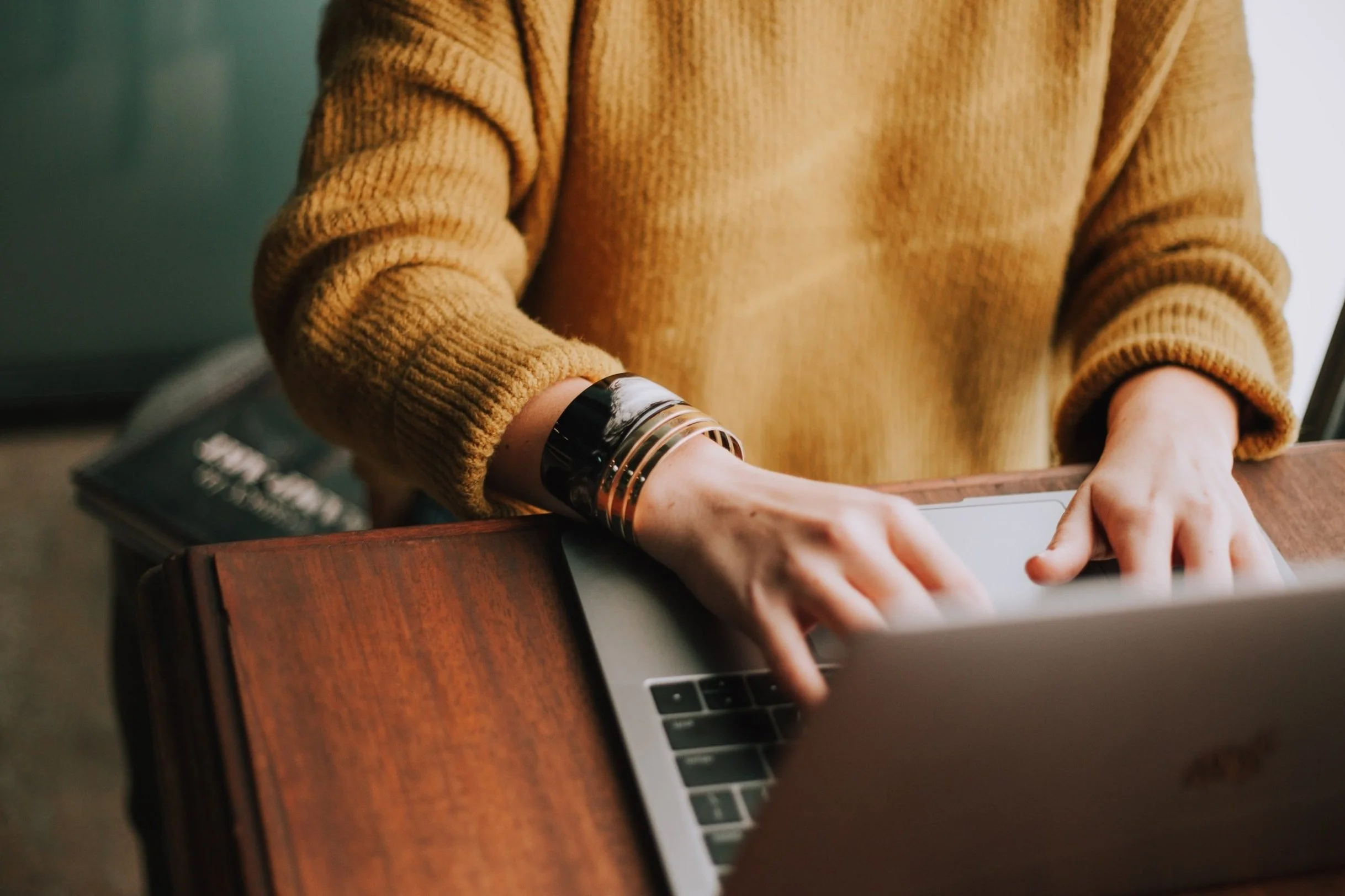 A person wearing a tan sweater typing on a laptop at a wooden table