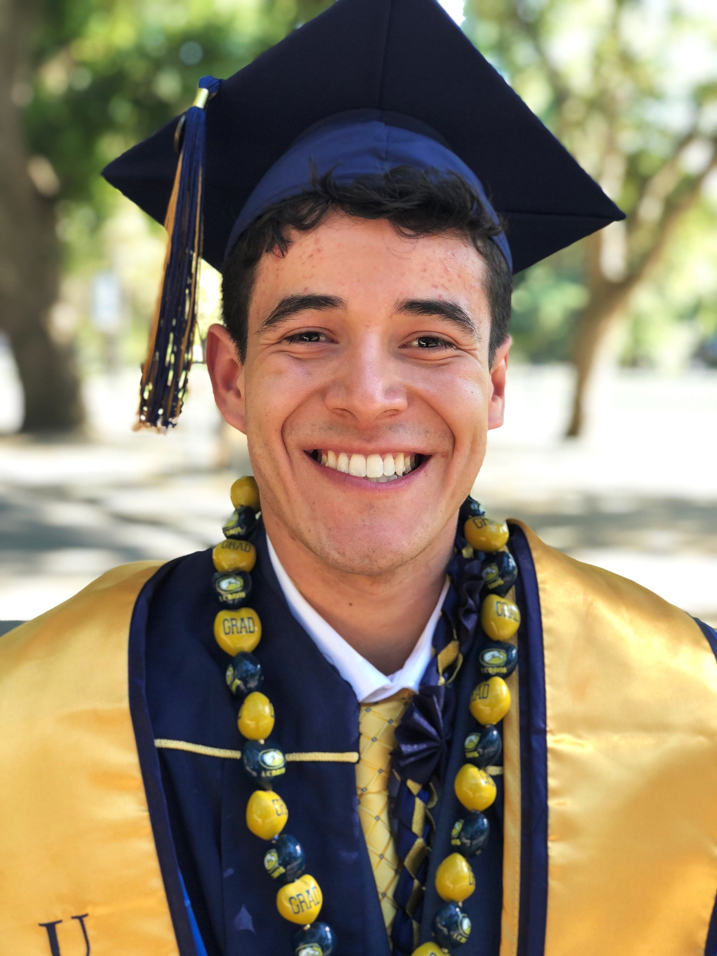 Young man smiling in graduation cap and gown, wearing a beaded necklace with yellow and blue beads and a yellow tie, outdoors with green trees in the background.