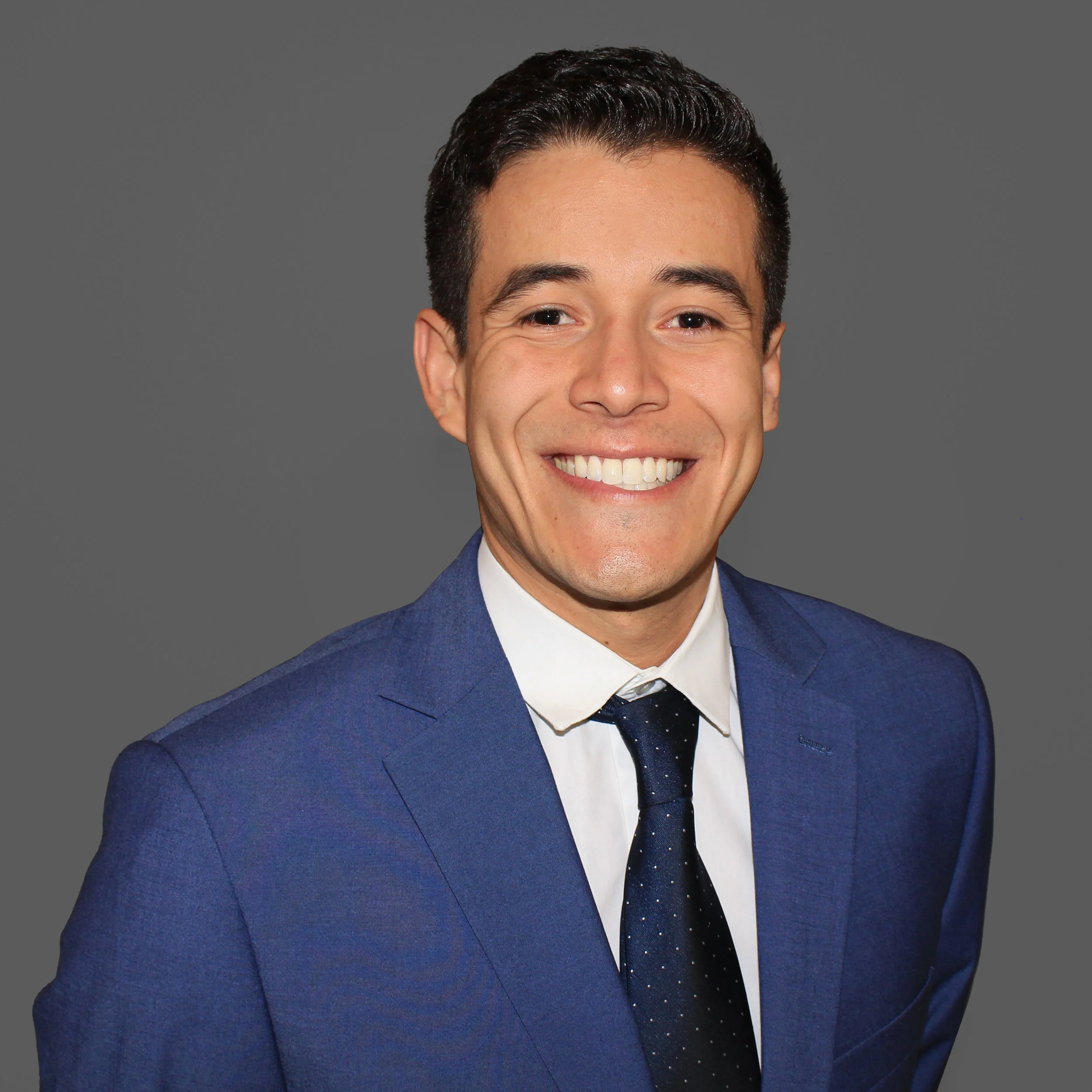 A smiling man in a blue suit, white shirt, and dark tie against a gray background.