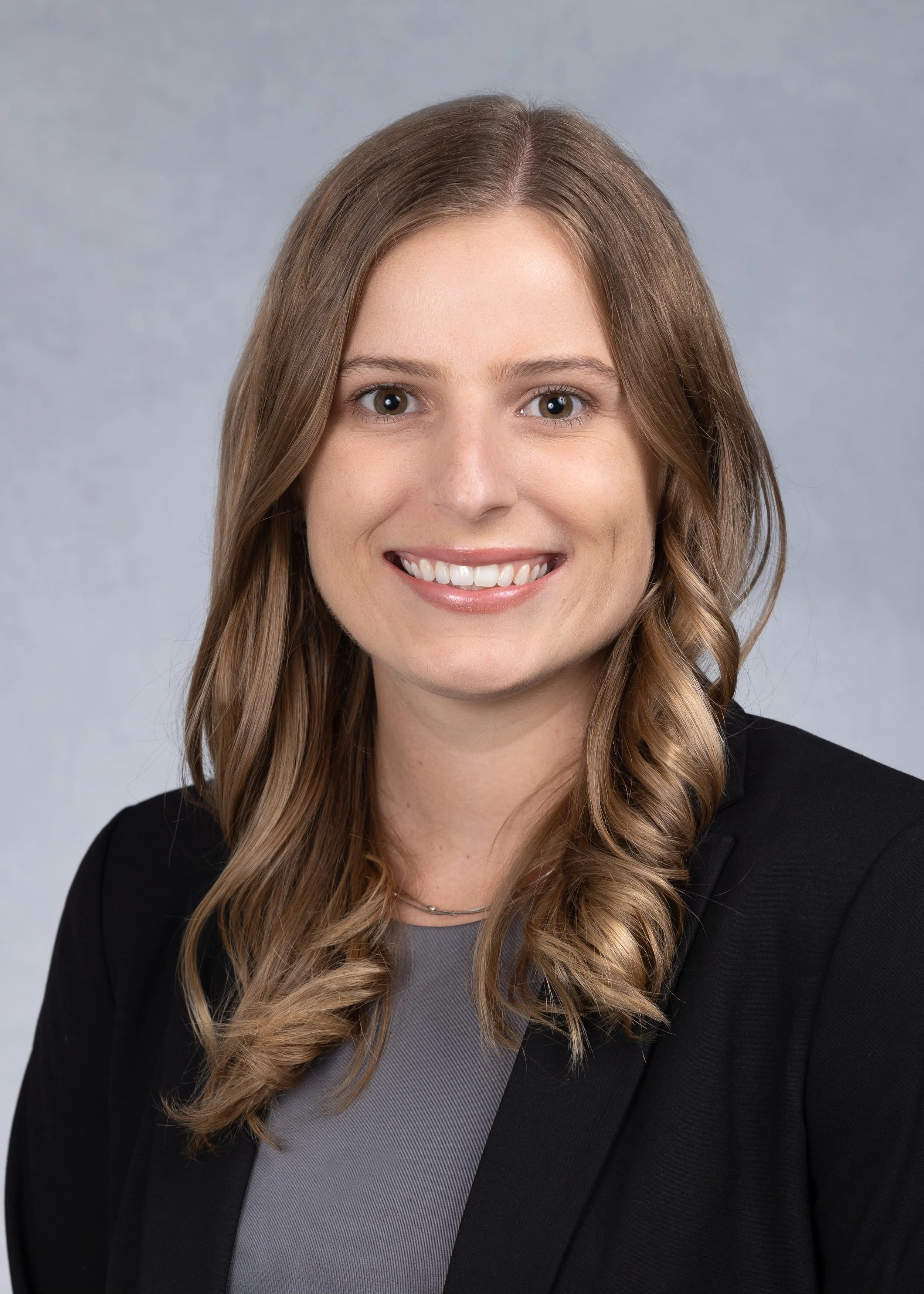Professional woman with wavy, shoulder-length brown hair, wearing a black blazer and a gray top, smiling against a neutral gray background.