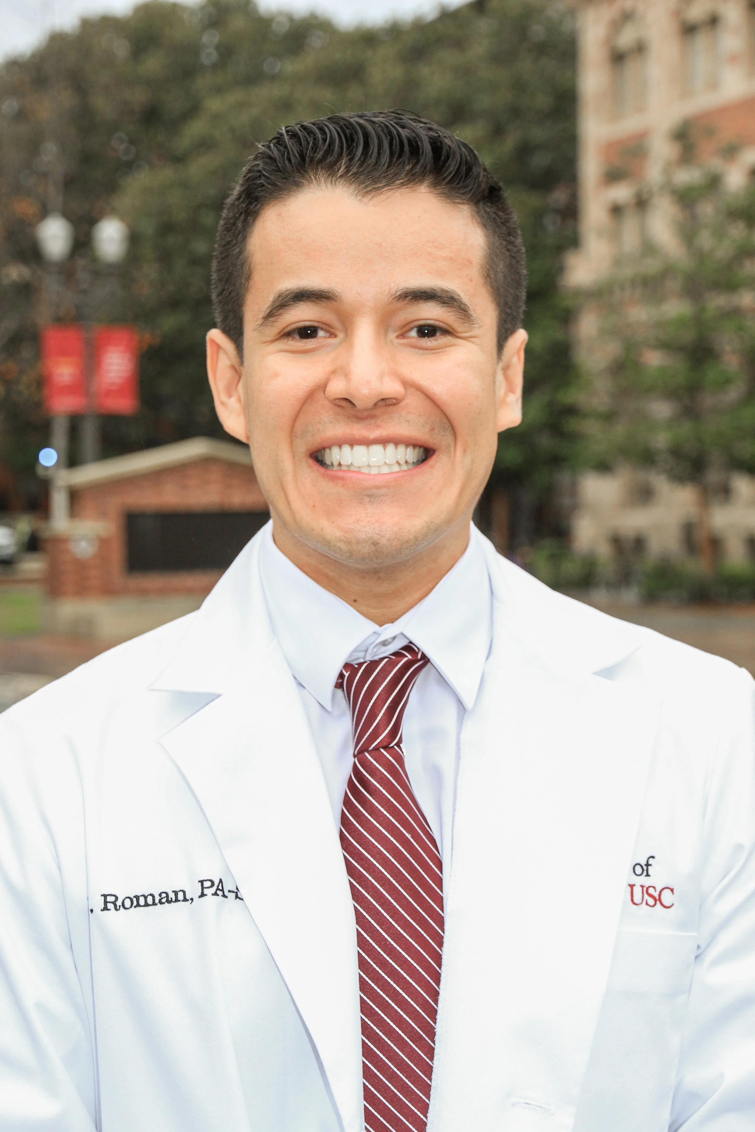 A smiling man in a white lab coat with 'Roman, PA-C' embroidered on it, standing outdoors with trees and a building in the background.