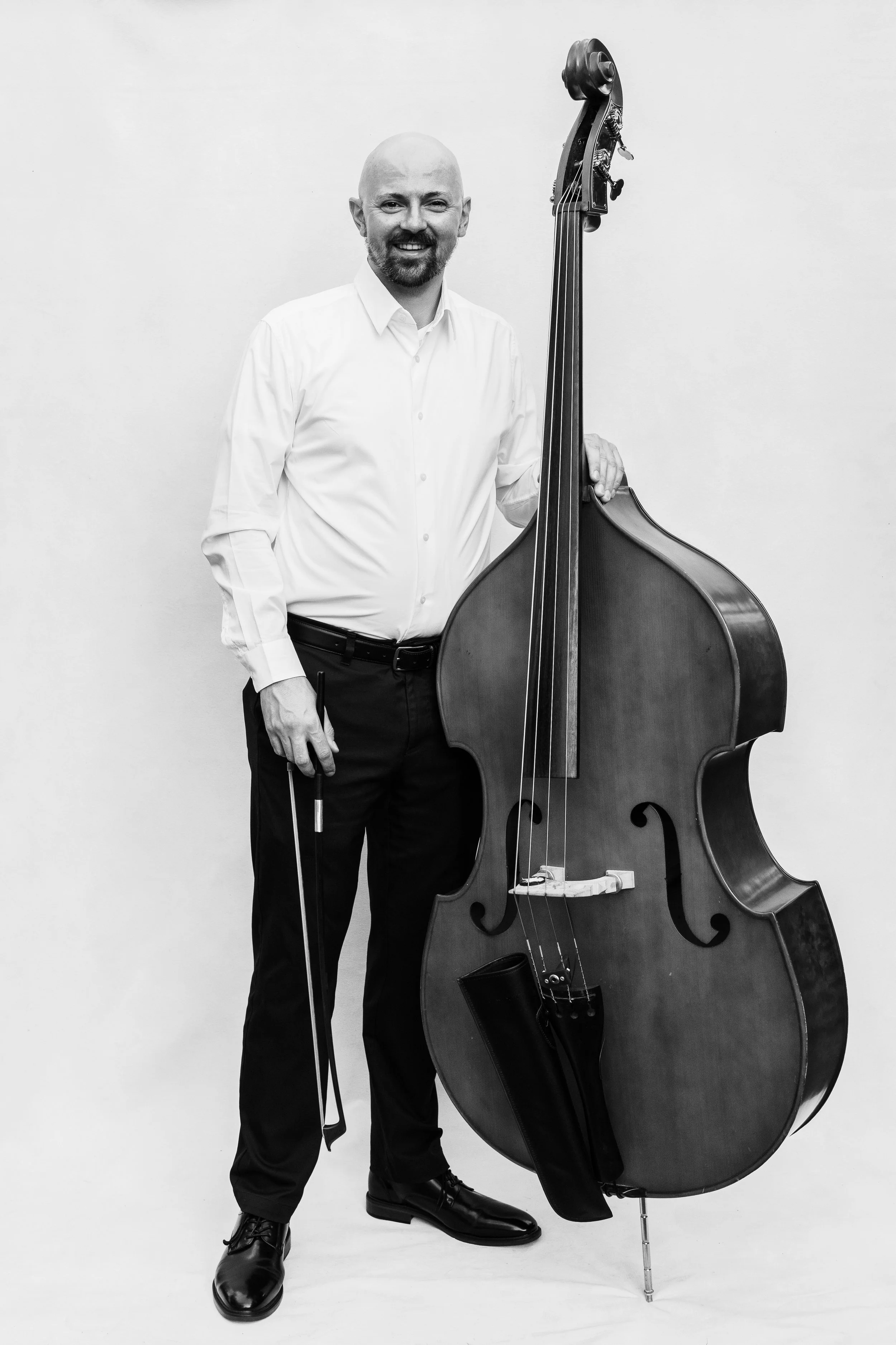 Adam Willey, a bald man with beard in dress shirt holding a double bass and bow, standing against a plain light-coloured background.
