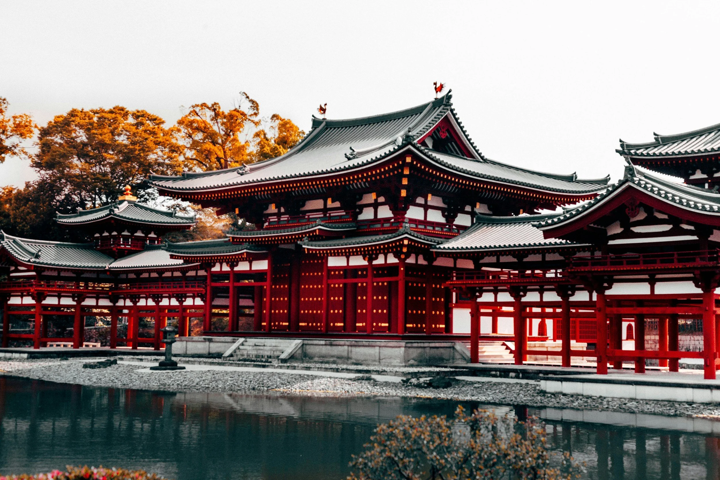 Traditional Japanese temple with red and white wooden architecture surrounded by a pond and autumn trees in the background.