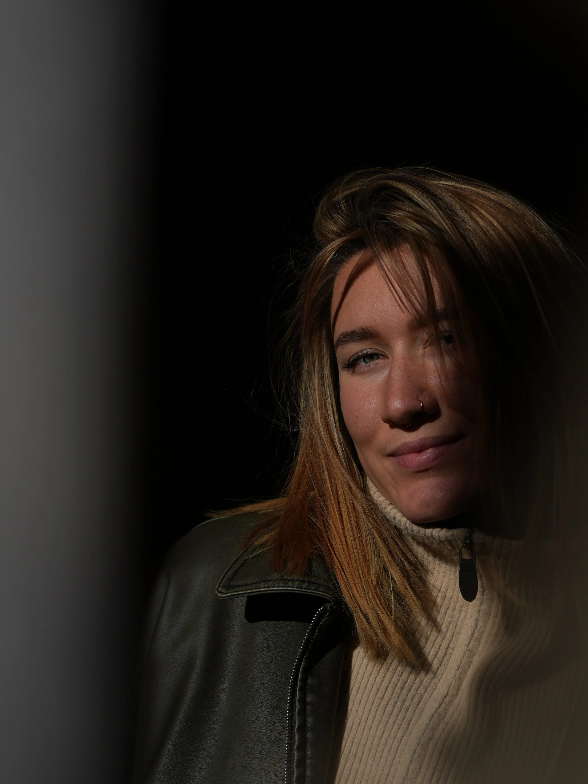 A young woman with shoulder-length brown hair, a nose ring, and a subtle smile, wearing a beige turtleneck and a dark jacket, illuminated on one side against a dark background.