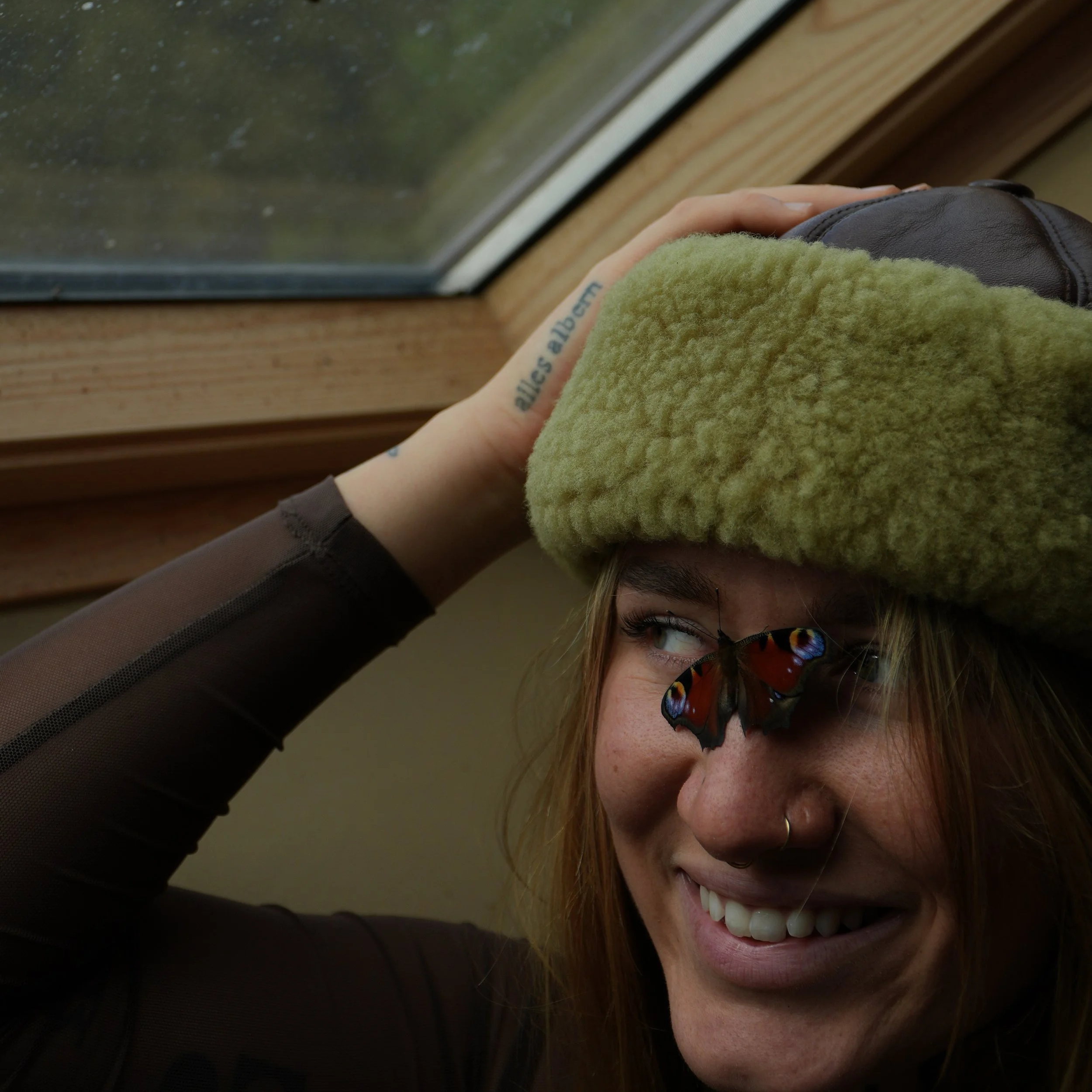 A person with a butterfly on their face, laughing and wearing a fuzzy hat, near a window.