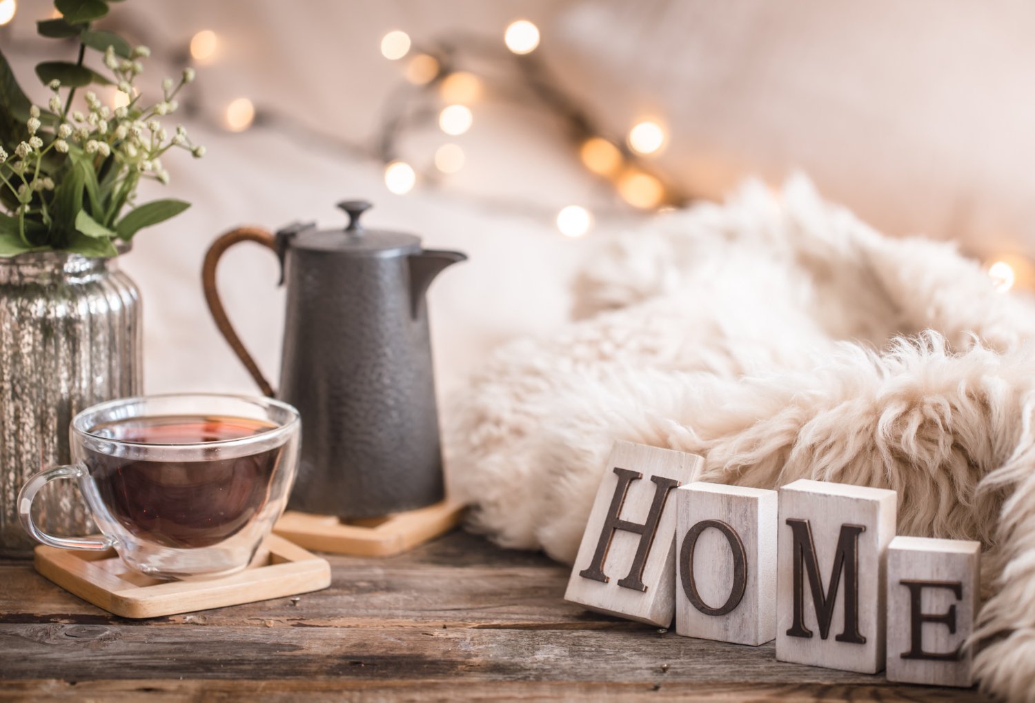 Cozy home scene with a glass teacup filled with tea, a dark gray teapot, a vase with green foliage, a fluffy blanket, and wooden blocks spelling 'HOME,' decorated with soft warm lights.