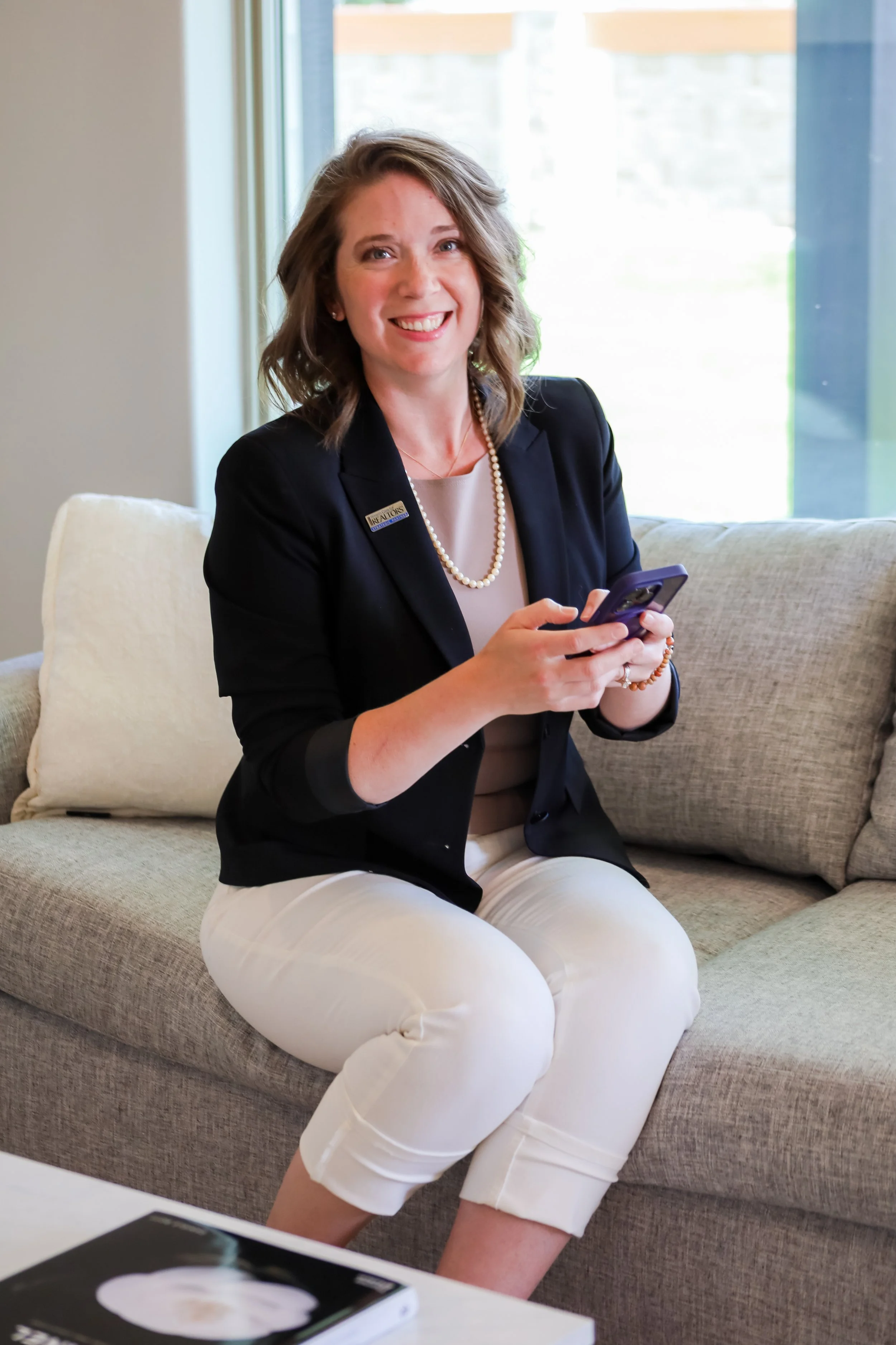 A woman with shoulder-length curly hair, wearing a black blazer, a pearl necklace, and white pants, is sitting on a beige sofa holding a smartphone and smiling. She has a name badge on her blazer.