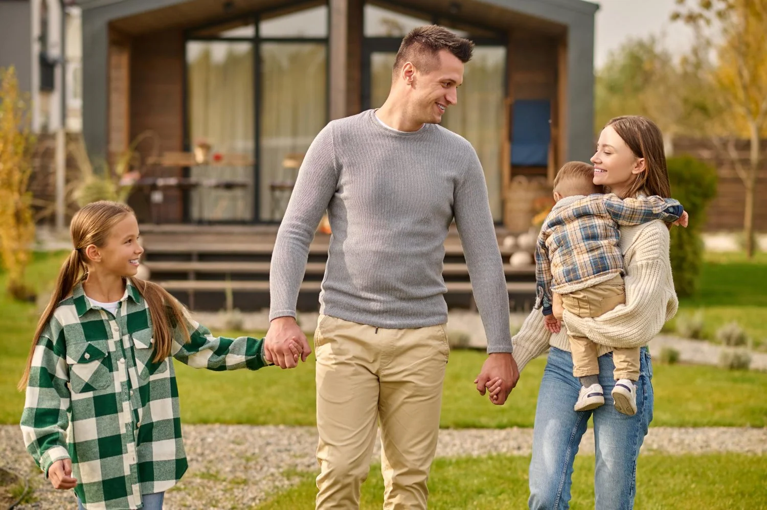 A family of four holding hands and smiling outdoors in front of a modern house with a deck, during fall.