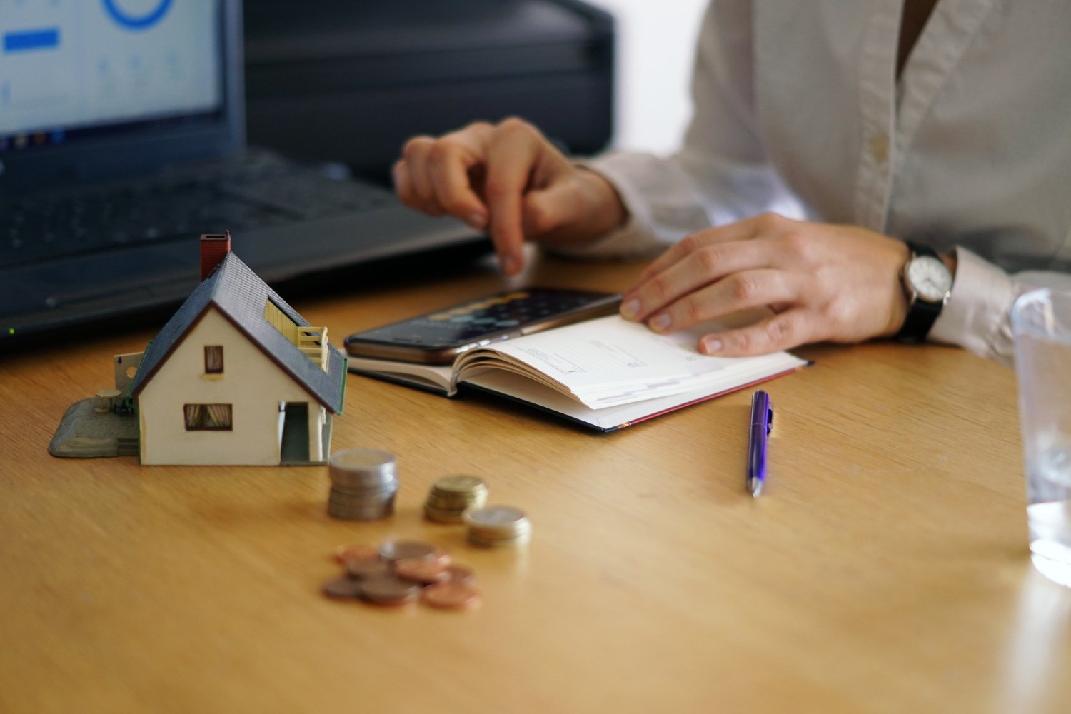 Person working at a desk with a miniature house model, coins, and a notebook, with a laptop and smartphone.