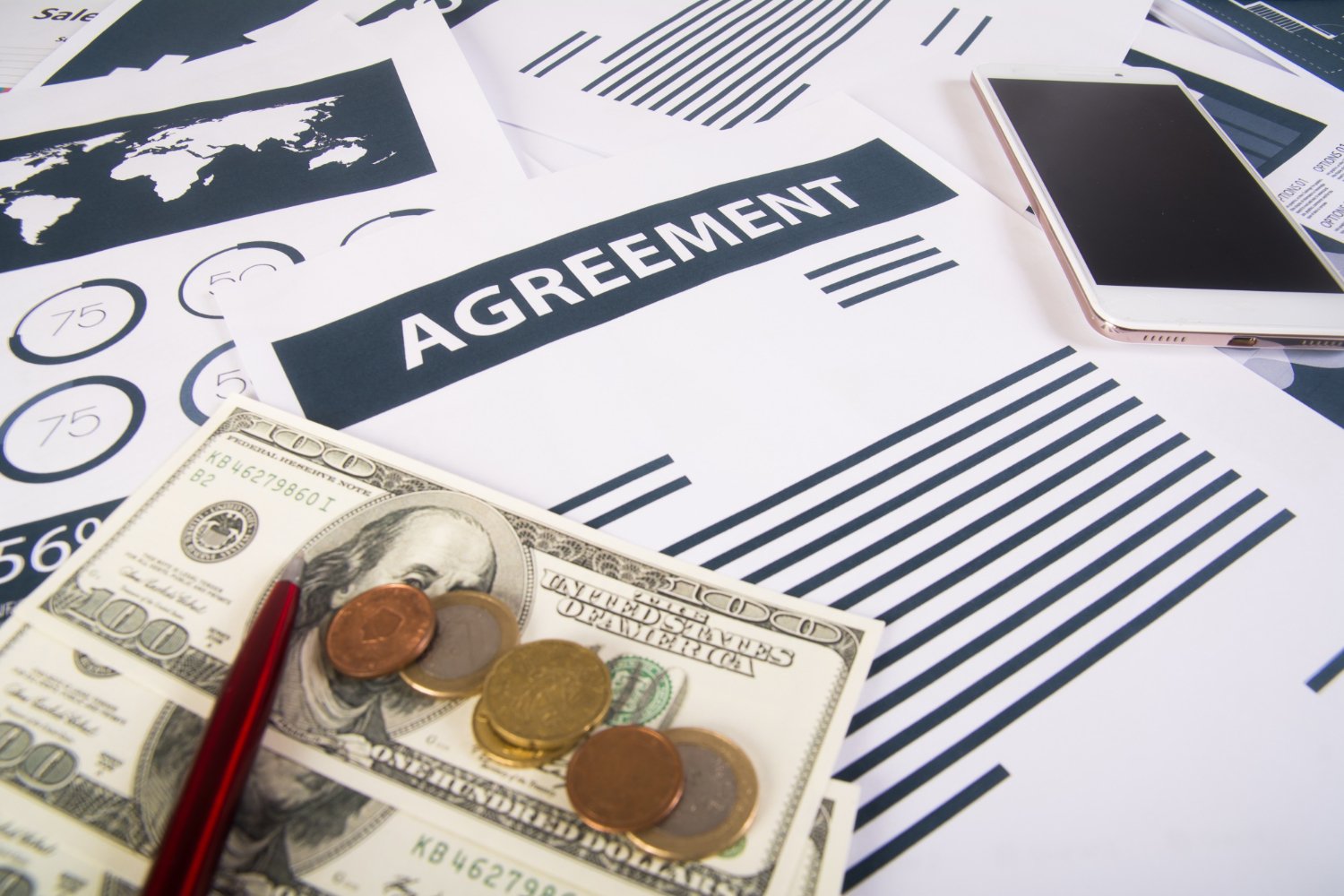 Financial documents including a contract labeled 'AGREEMENT', a US hundred-dollar bill, coins, a red pen, and a smartphone on a desk.