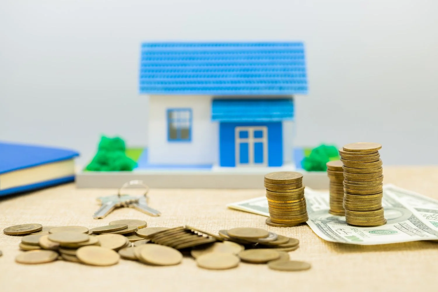 Stacks of coins and dollar bills in front of a miniature blue and white house model, with a set of keys and a blue book nearby.