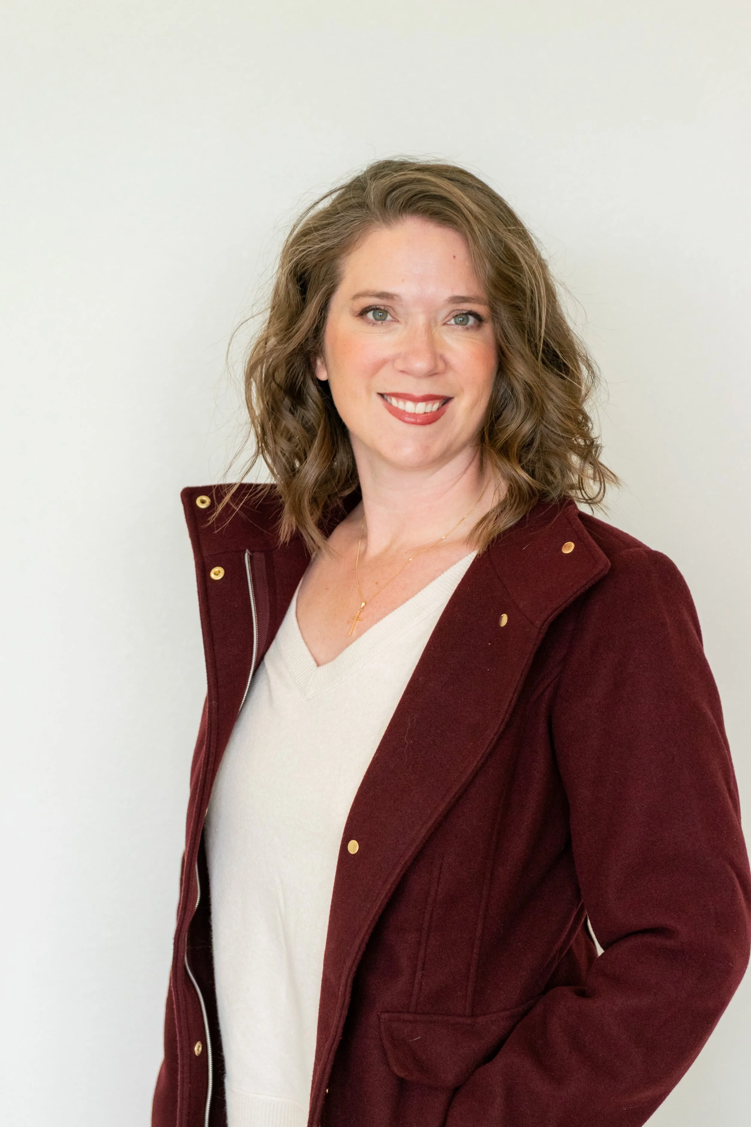 Portrait of a smiling woman with wavy brown hair, wearing a burgundy jacket over a cream sweater, standing against a plain white background.