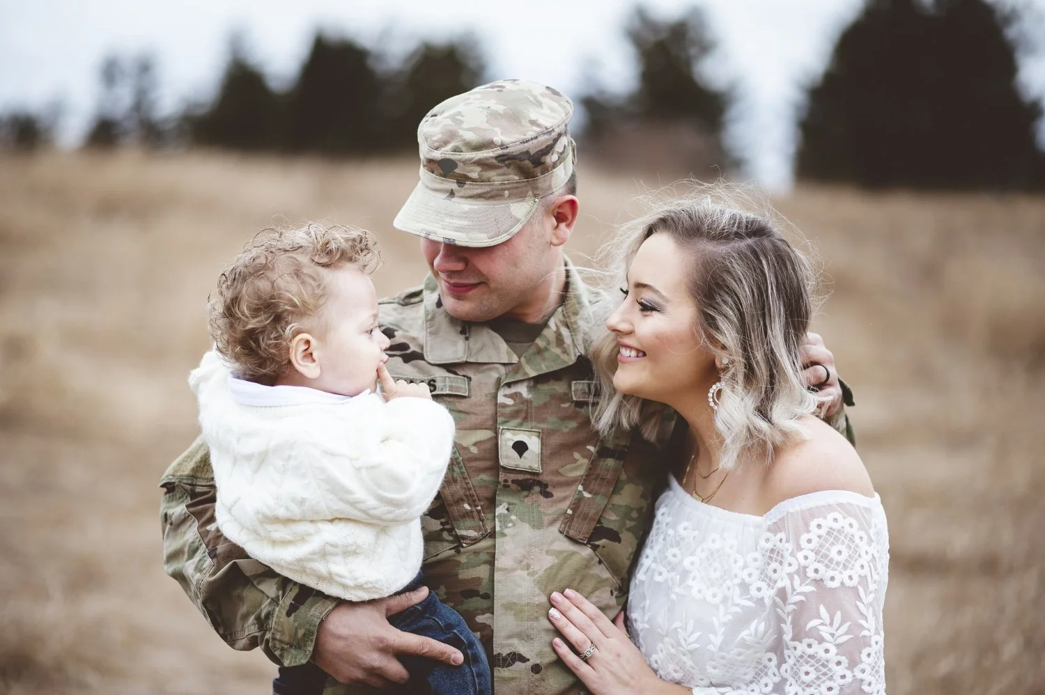 A soldier in uniform holding a young child with curly hair, while a woman with wavy hair and earrings stands close, smiling, in an outdoor field with trees in the background.