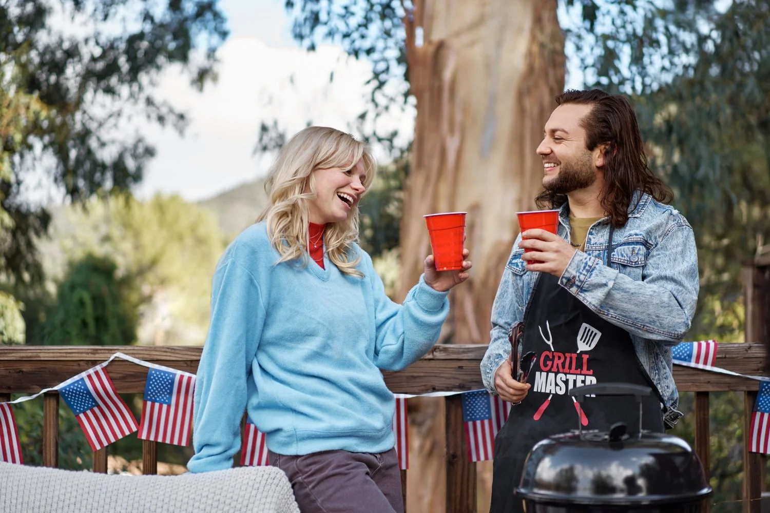 Two people celebrating outdoors with red plastic cups, on a deck decorated with American flags, one person is wearing a "Grill Master" apron, and a grill is visible.