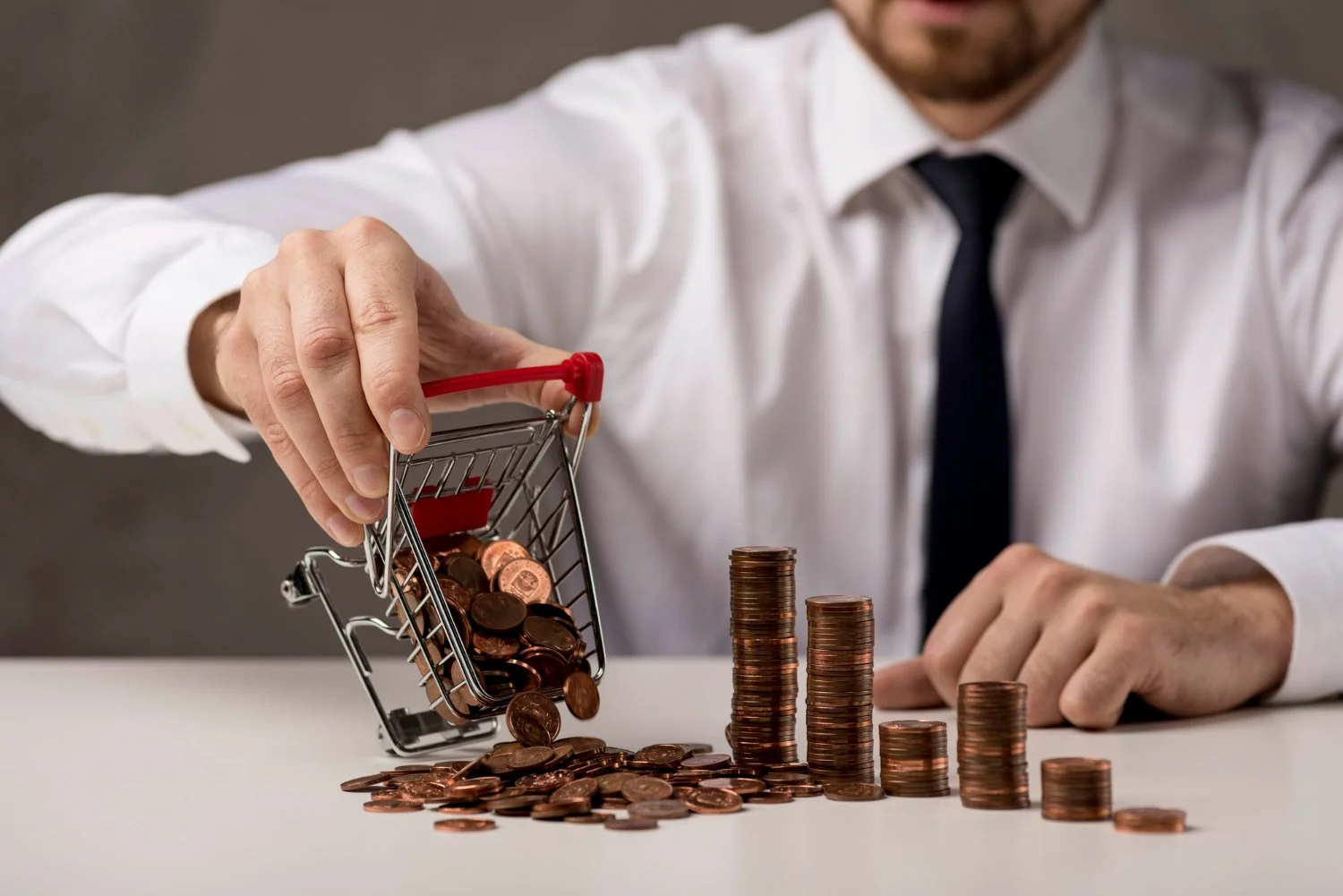 A man in a white shirt and black tie is pouring coins from a small shopping cart onto a table with stacked coins arranged in ascending order.